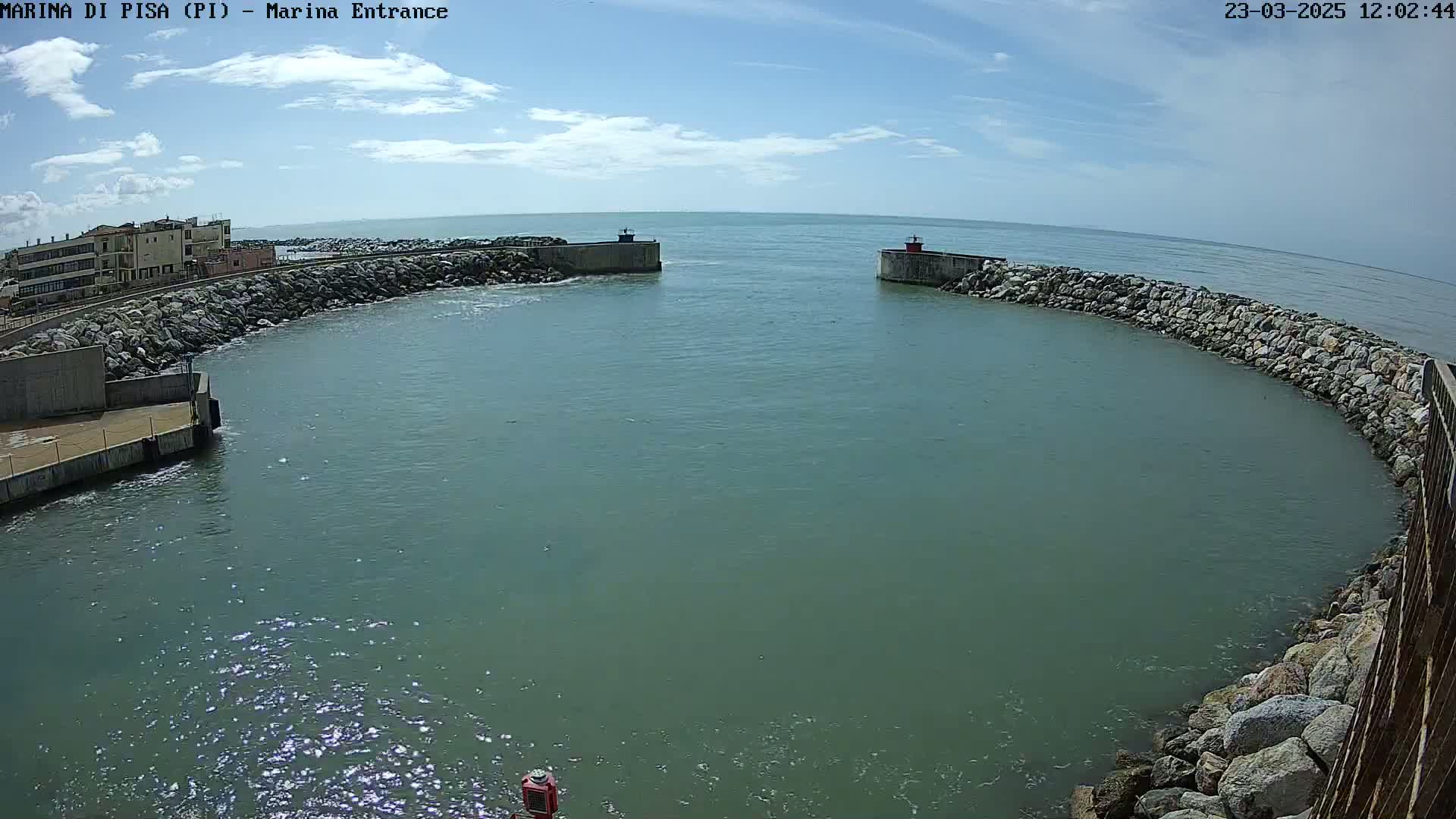 A harbor entrance with calm, greenish-blue water is surrounded by rock walls under a partly cloudy sky.