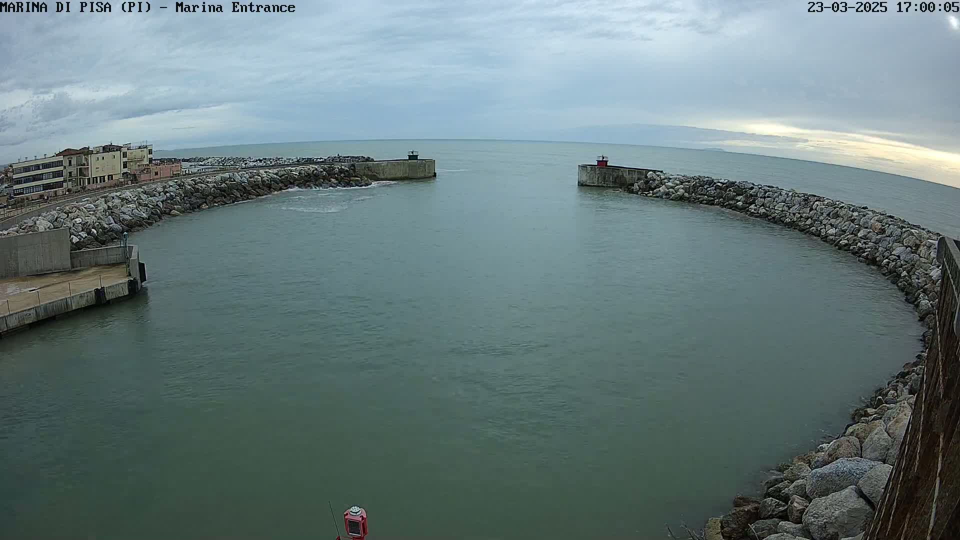 A harbor entrance with calm, gray-green water is surrounded by rock walls, with buildings and a cloudy sky visible in the background.