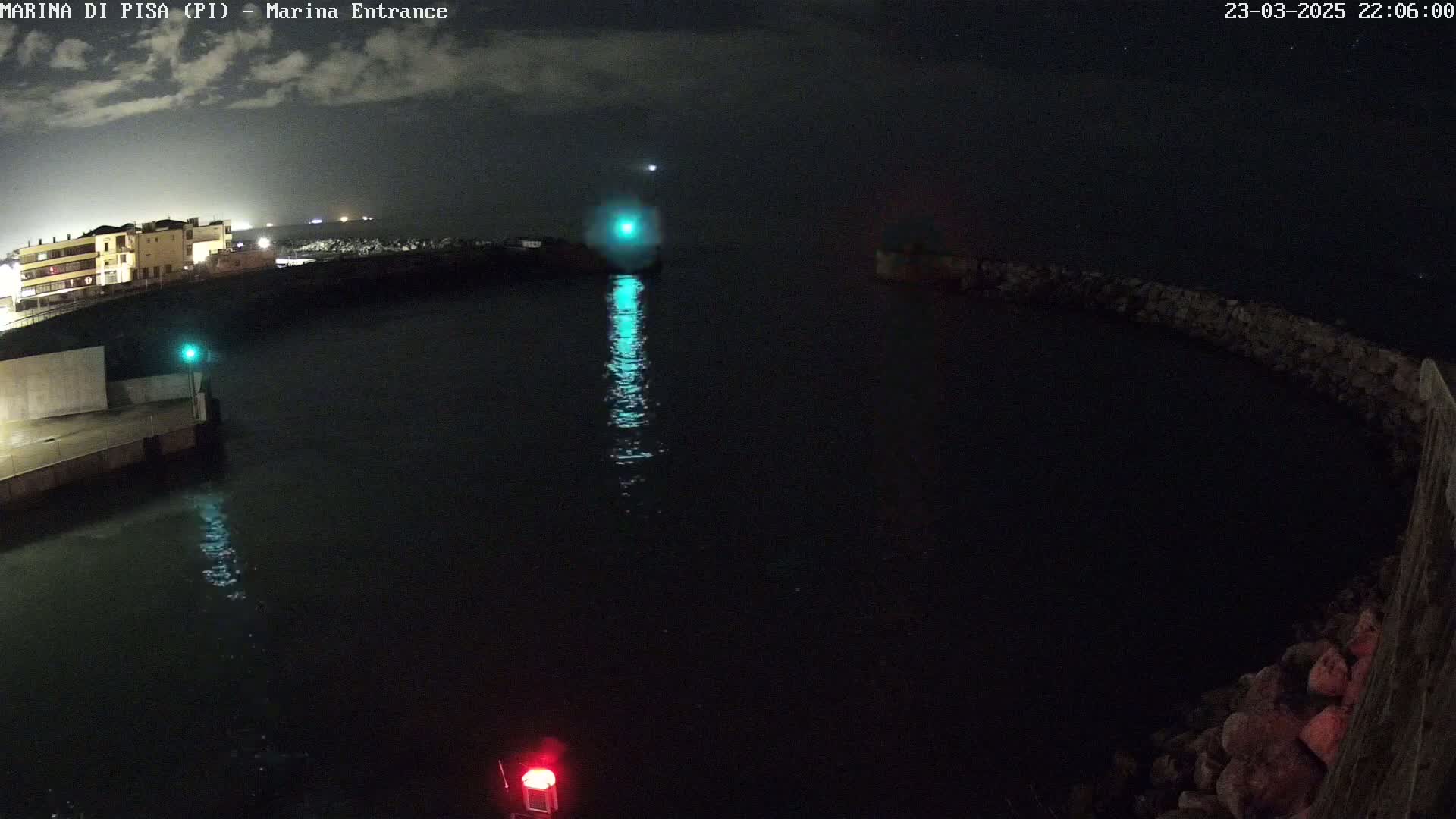 A nighttime view of a dark harbor with a building in the background, calm water reflecting lights, and a rock wall.