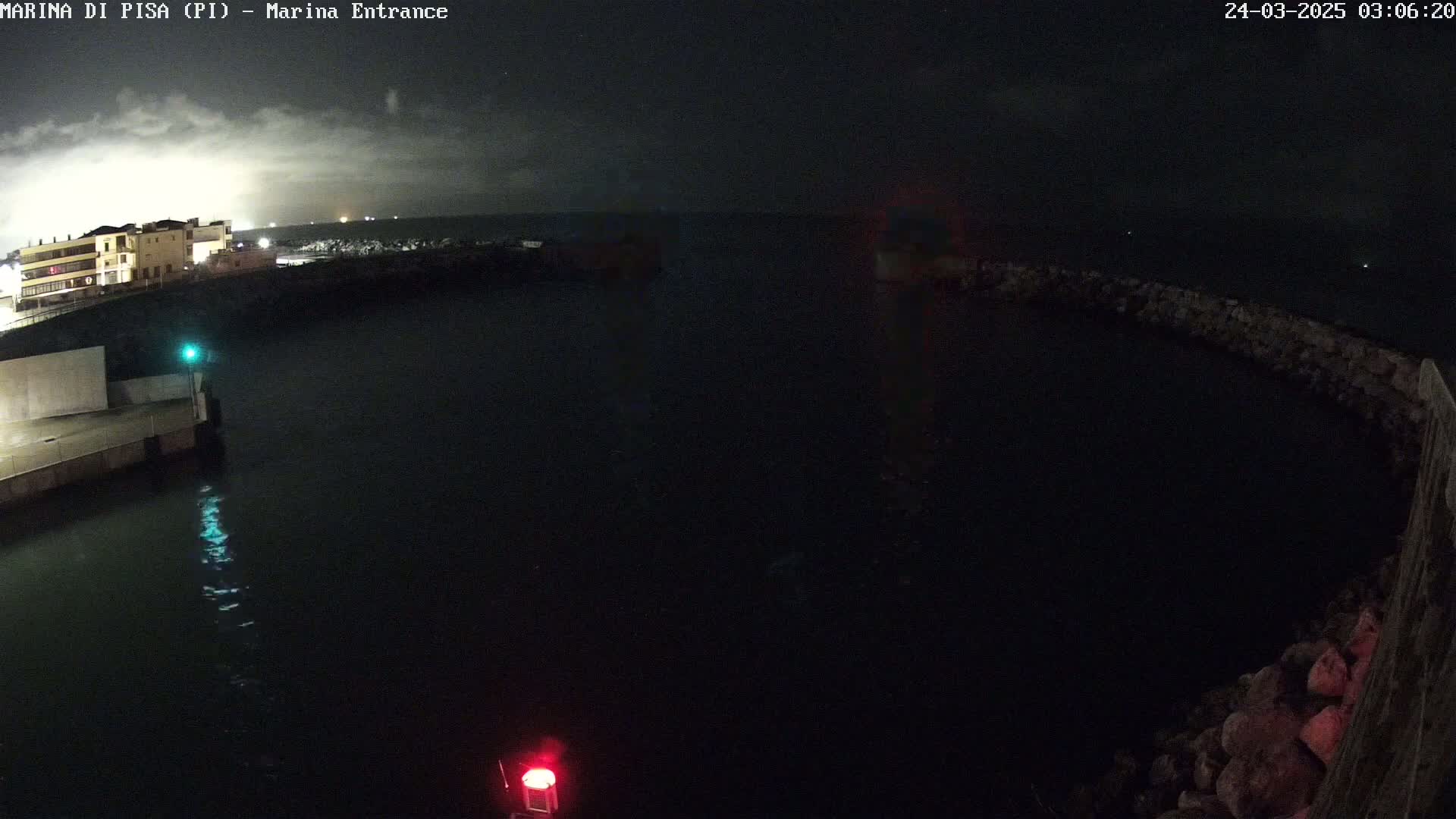 A nighttime view of a dark harbor with calm water, a building in the distance, and a partially visible stone wall.