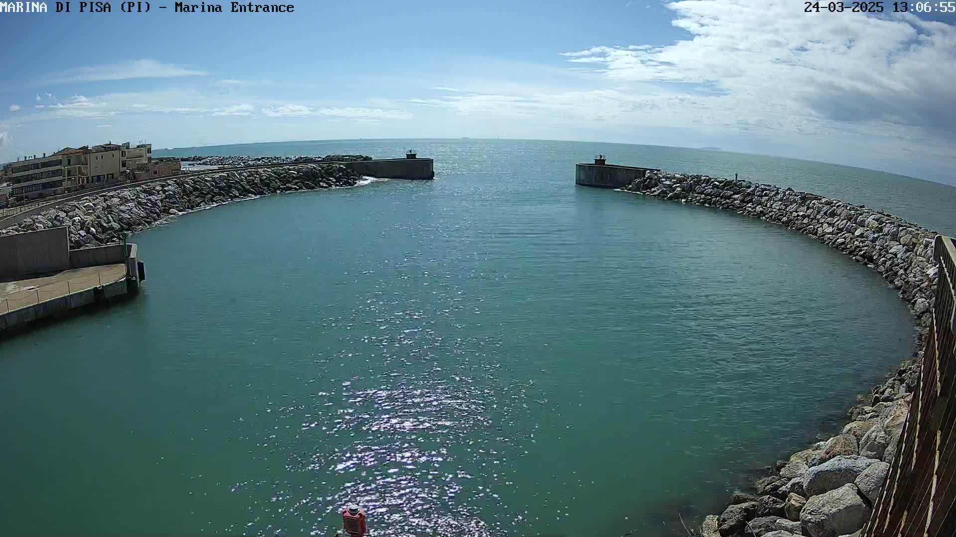 A marina entrance with calm, sparkling teal water is nestled between two rocky breakwaters under a mostly sunny sky with some clouds.