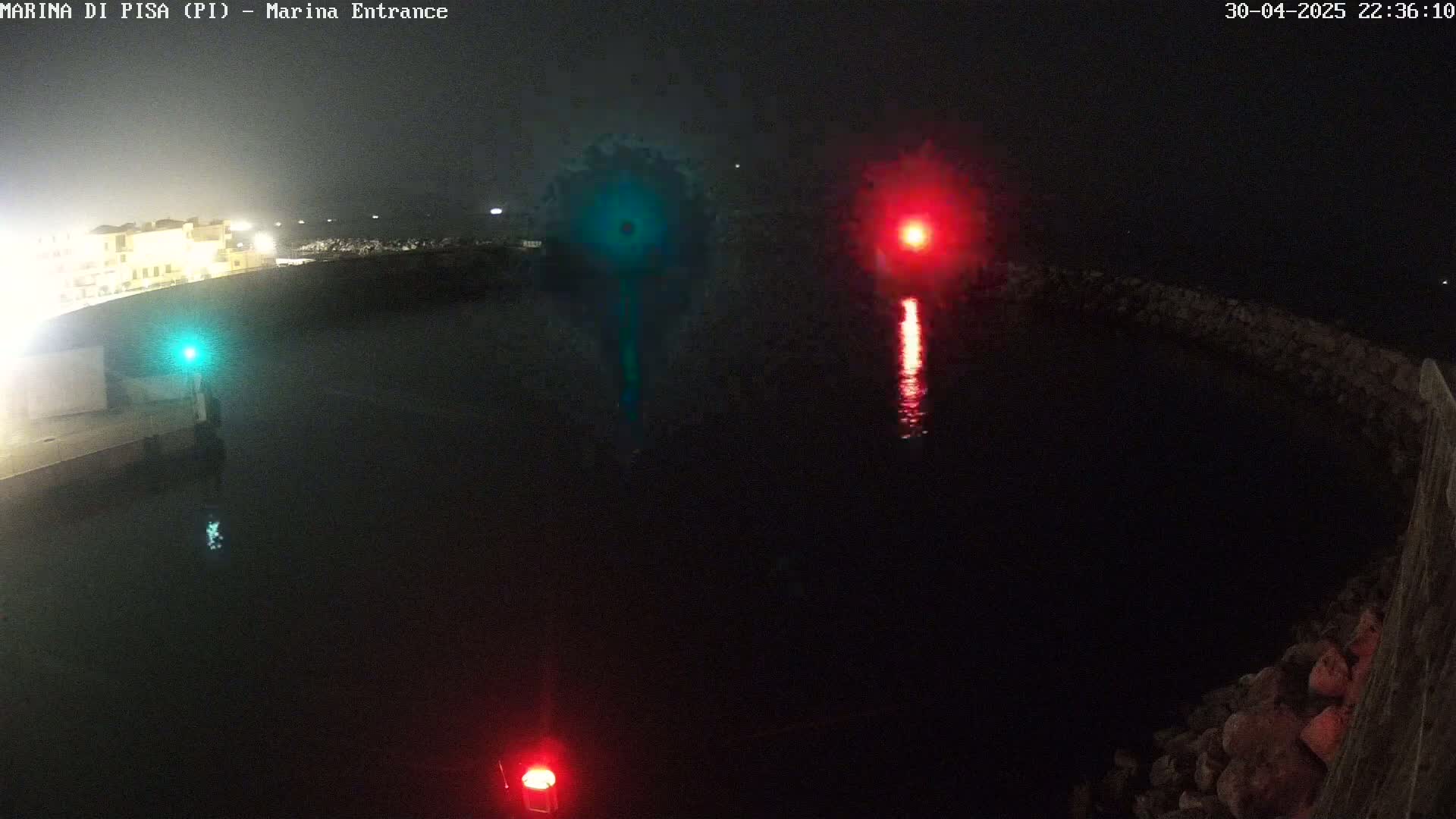 At night, a dark body of water is illuminated by a green and a red light, reflecting on its surface near a stone structure.