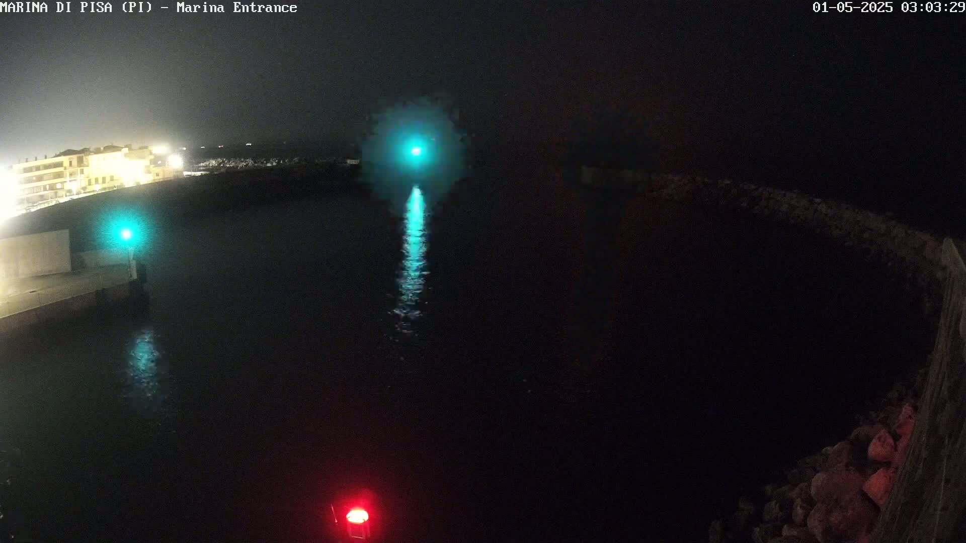 A nighttime, high-angle view of a dark harbor with a bright teal light reflecting on the water and illuminated buildings in the background.