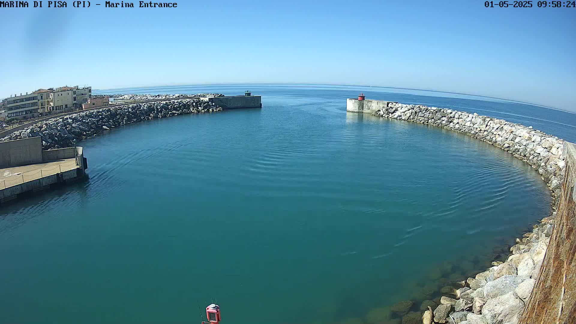 A marina entrance with teal water under a clear blue sky is surrounded by rock walls and a small building.