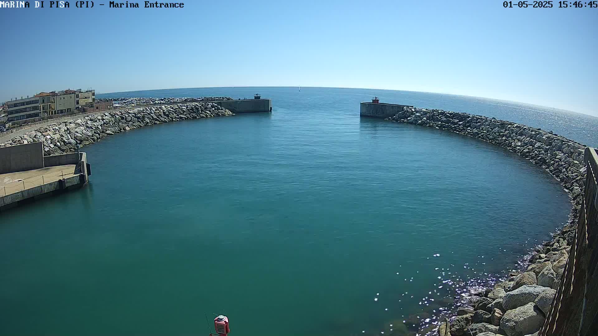 A marina entrance with calm, teal water under a clear blue sky is surrounded by rock walls and a building in the distance.