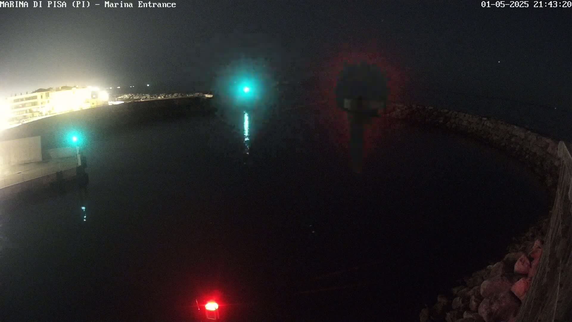 A nighttime view of a dark harbor with illuminated buildings in the distance, harbor lights, and a dark, rocky breakwater.
