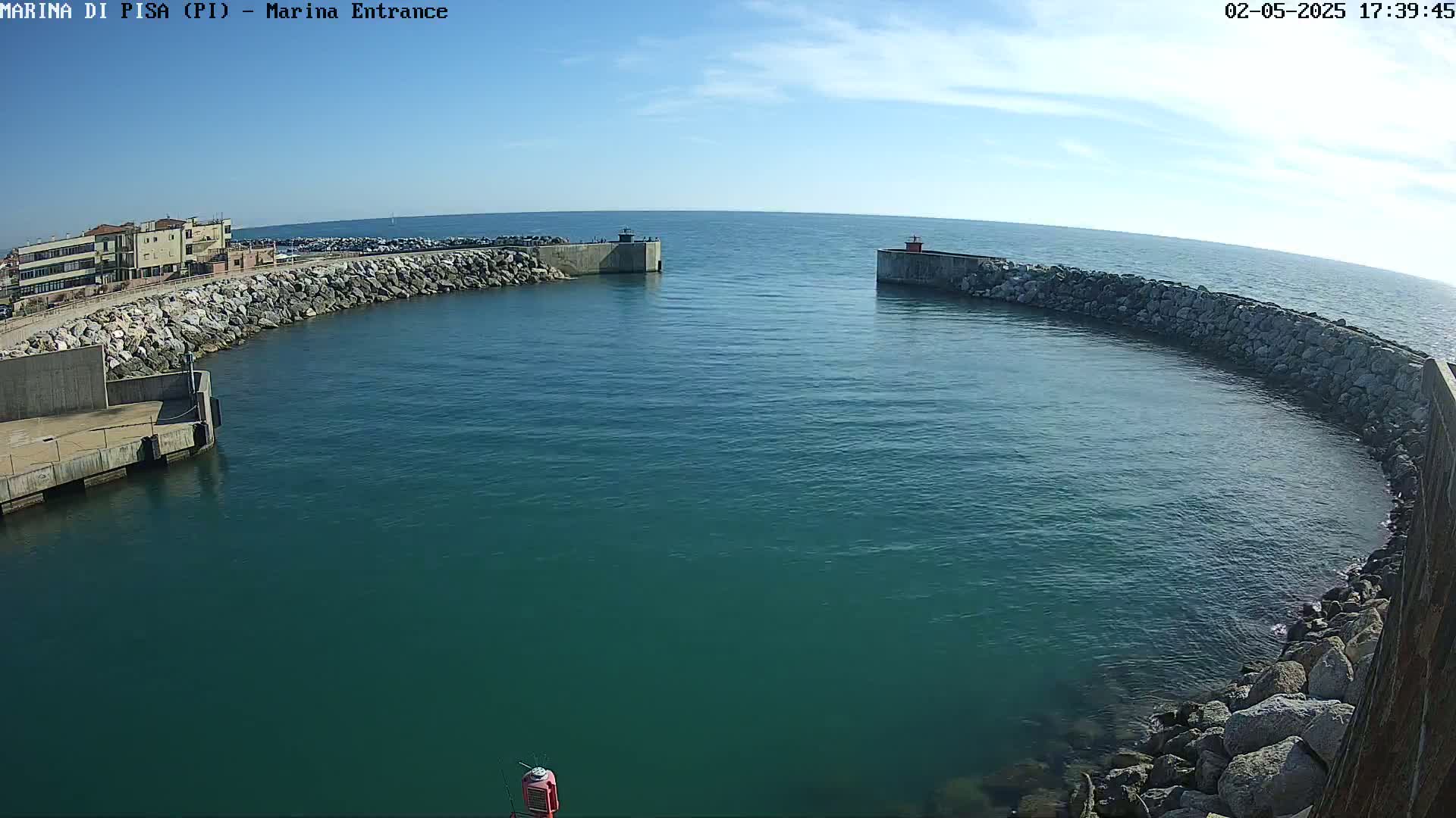 A marina entrance with calm, teal water under a mostly clear sky is flanked by rock walls and a building.