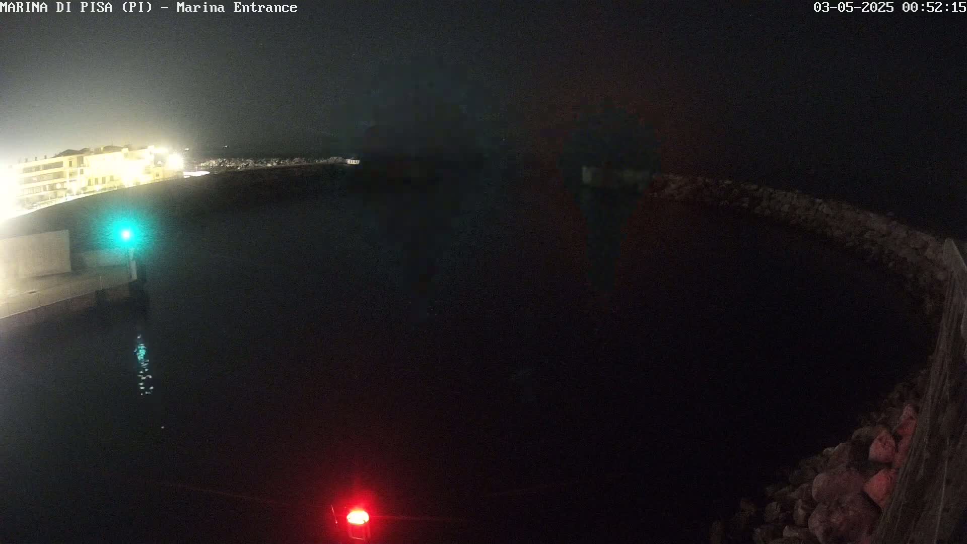 A nighttime, dark view of a marina entrance with a body of dark water, illuminated by a green light on one side and a red light on the other, and buildings faintly visible in the background.