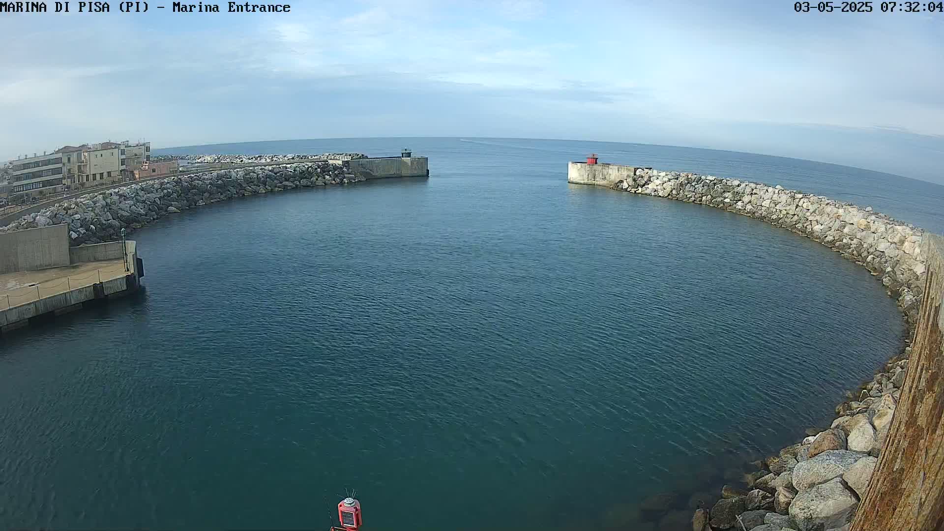 A calm, blue body of water is enclosed by a curved stone wall, with a small opening leading to a larger expanse of water under a mostly clear sky.