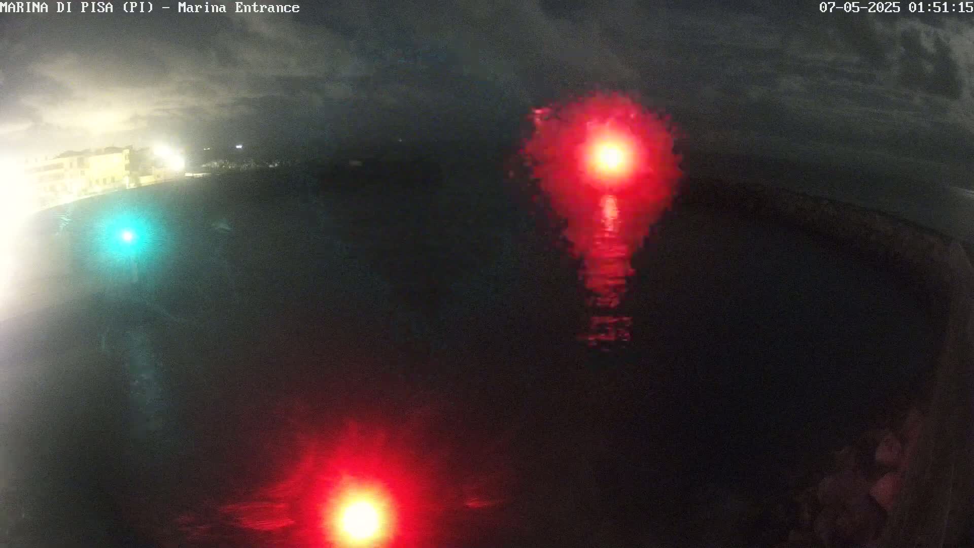 At night, under a cloudy sky, red and green lights are reflected on dark water near a shoreline.