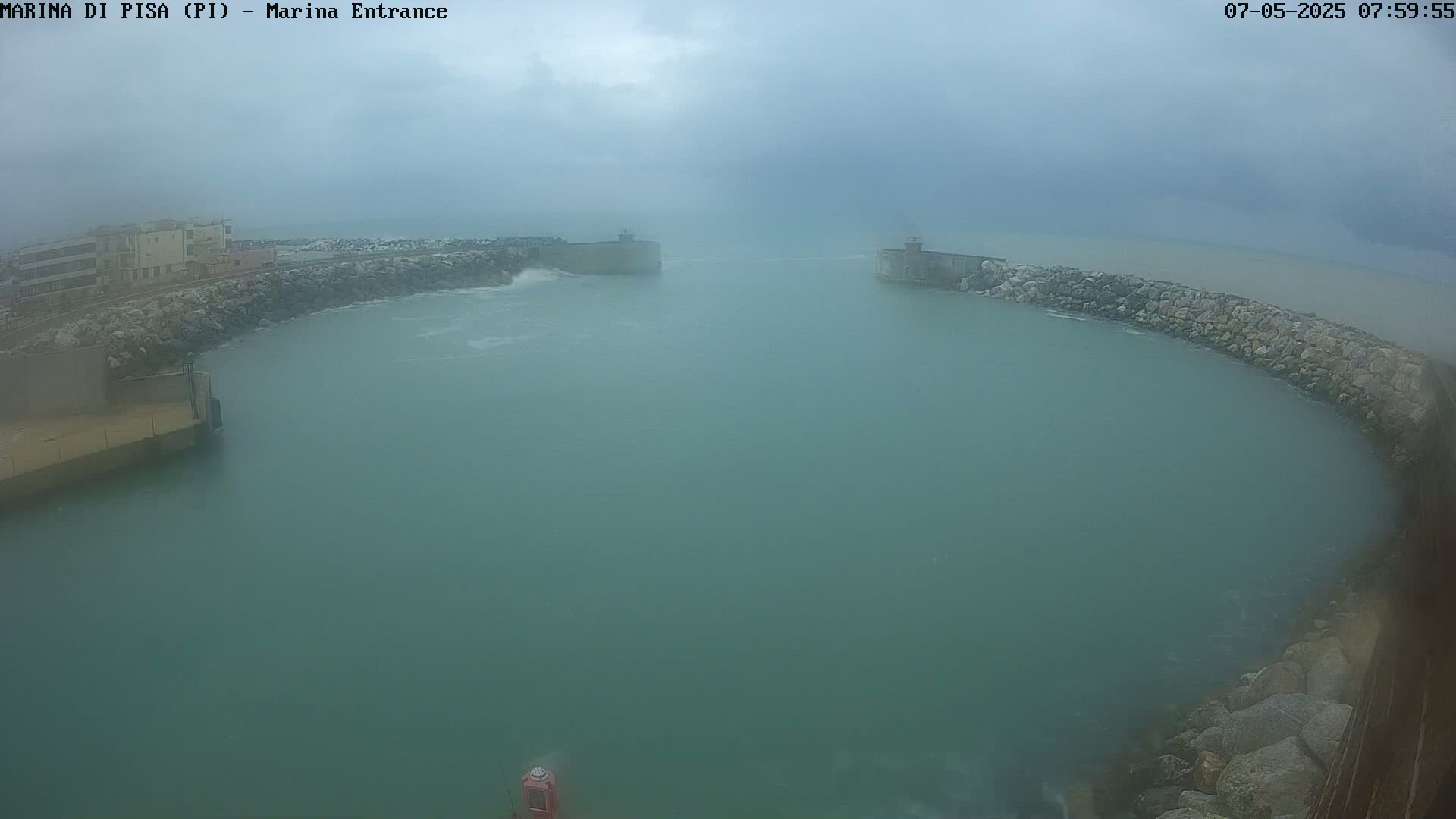 A hazy, overcast day reveals a harbor entrance with calm water partially enclosed by stone walls and small buildings on the shoreline.