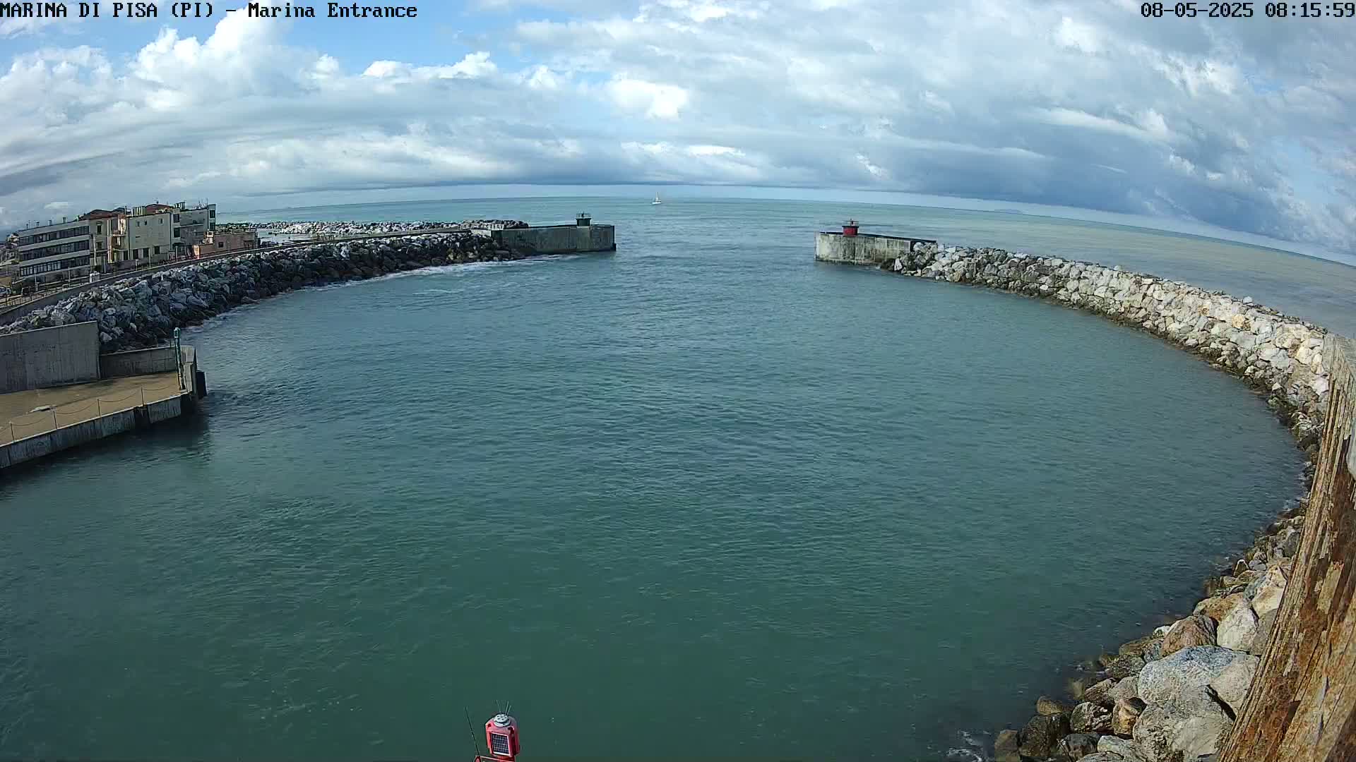 A partially cloudy sky overlooks a calm, teal-colored harbor enclosed by stone walls, with a small building visible in the distance.