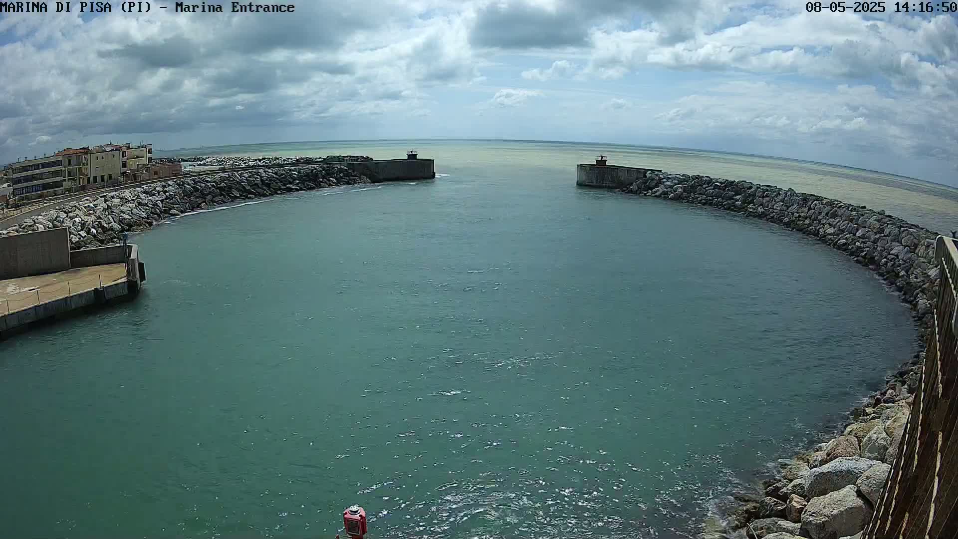 A calm, partly cloudy day shows a harbor entrance with a small building visible in the background, rock walls lining the waterway, and a mostly teal-colored body of water.