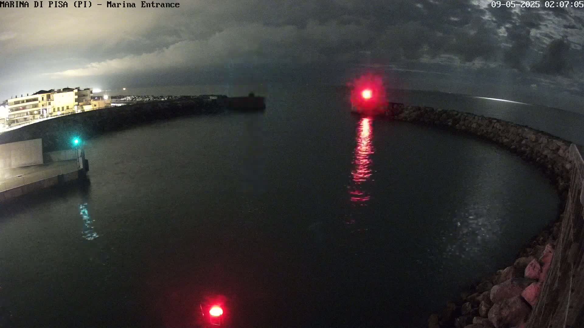 A nighttime view of a harbor entrance with calm water, illuminated by red and green navigation lights, under a cloudy sky.