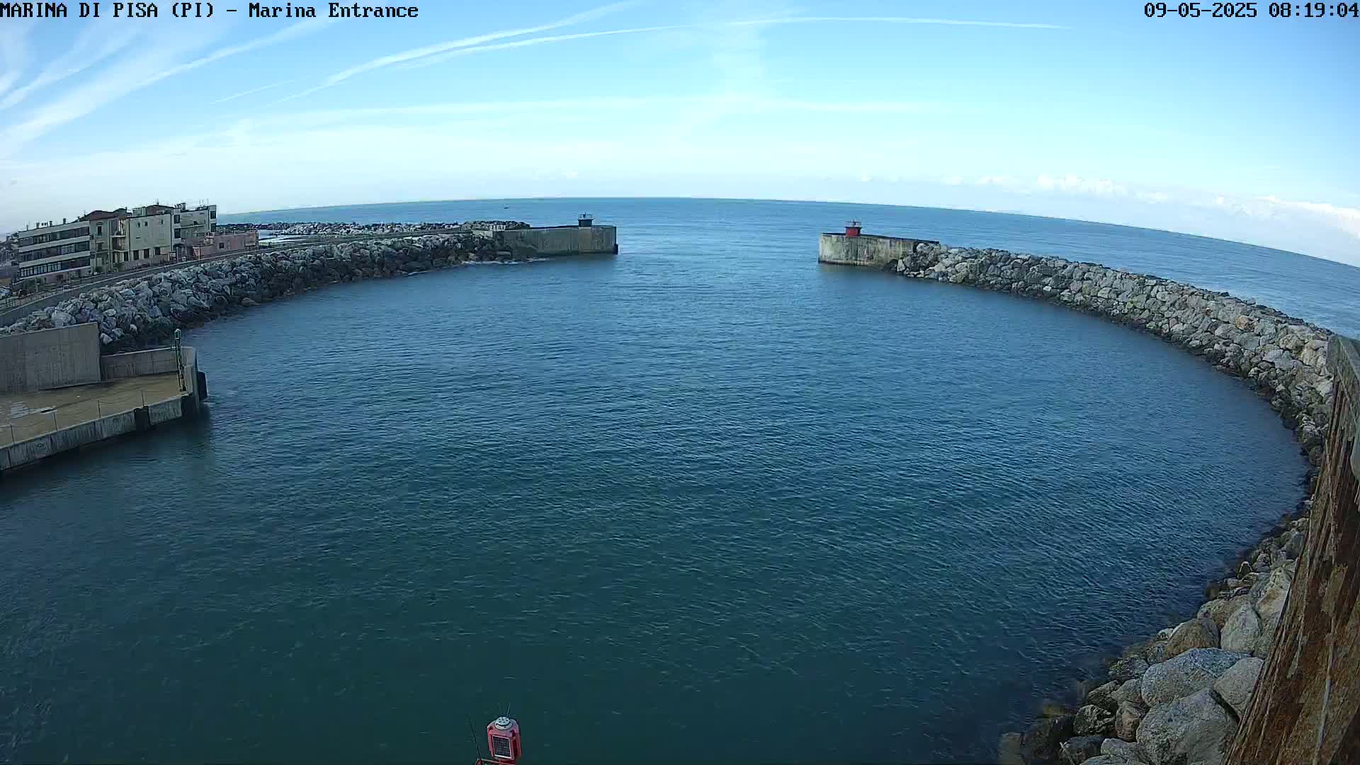A calm, blue body of water is partially enclosed by a curved rock wall, with a small building visible on the far shore under a clear, bright sky.