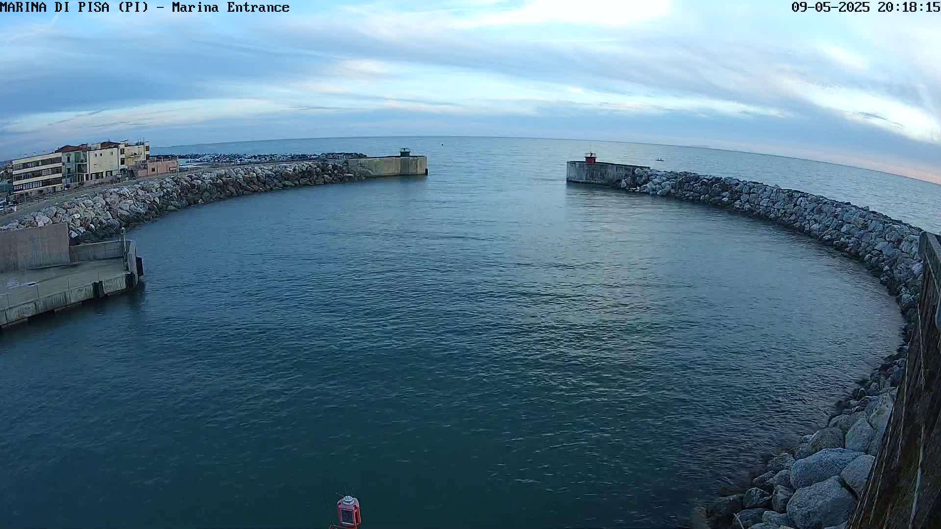 A calm, dark-blue body of water is nestled within a harbor, bordered by stone walls and buildings under a partly cloudy sky.