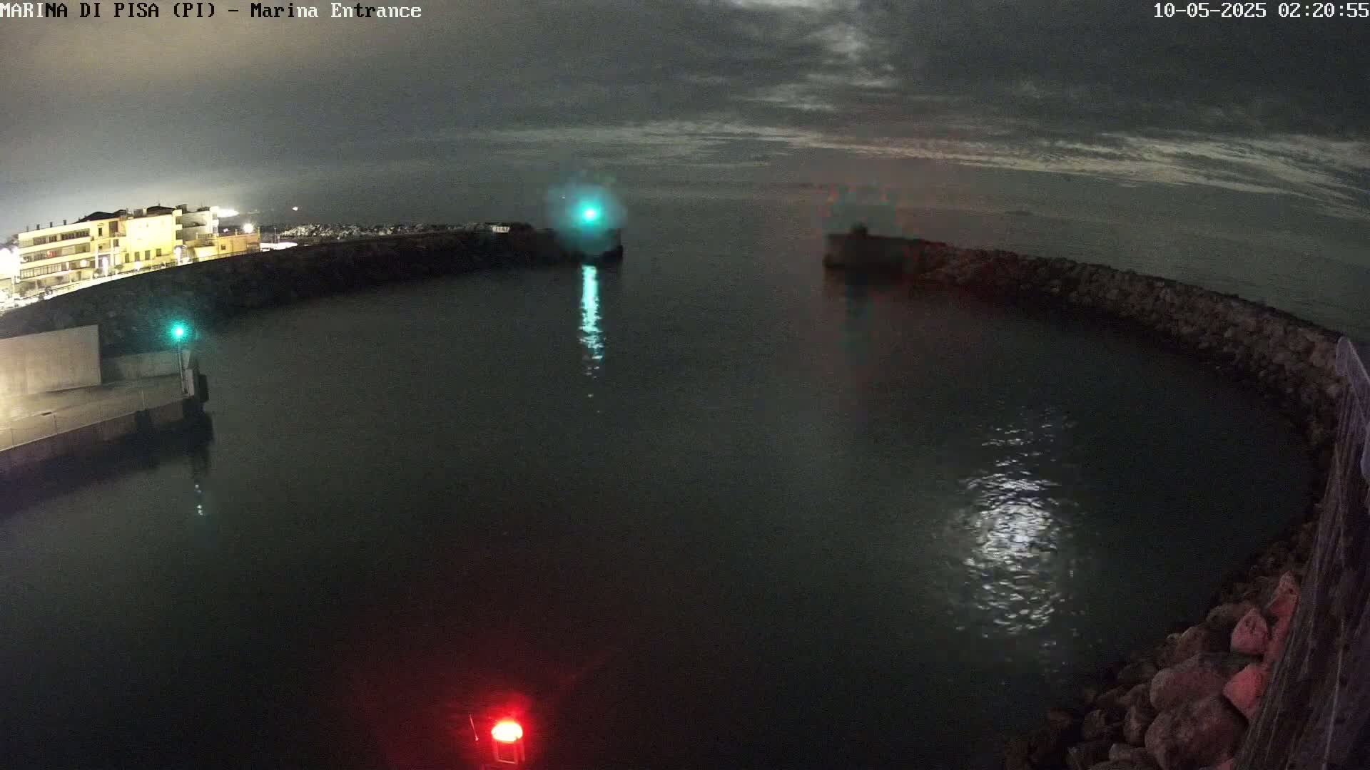 A nighttime view of a calm harbor, partially enclosed by a stone wall, with lights visible on the water and a building in the background under a cloudy sky.