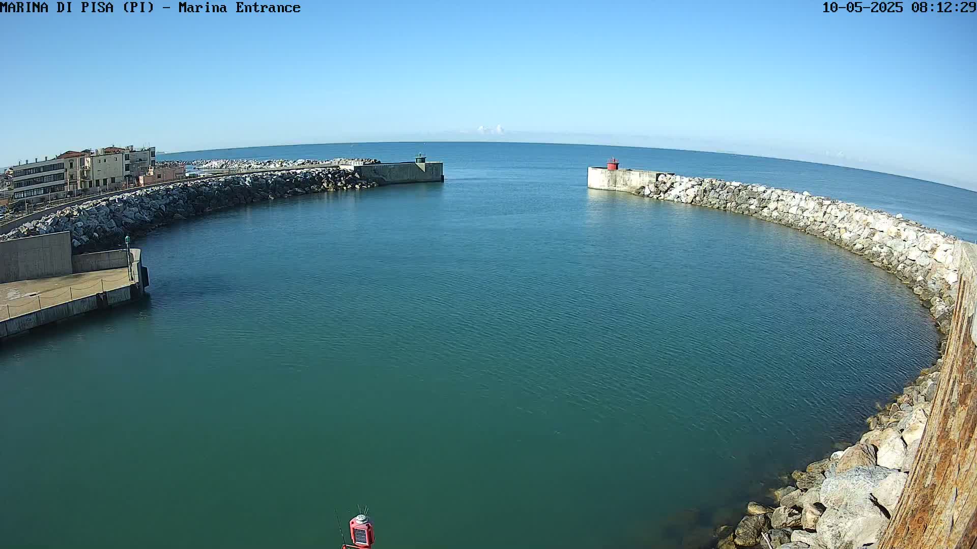 A calm, clear day reveals a harbor entrance with stone walls, a small building on one side, and the open sea in the distance.
