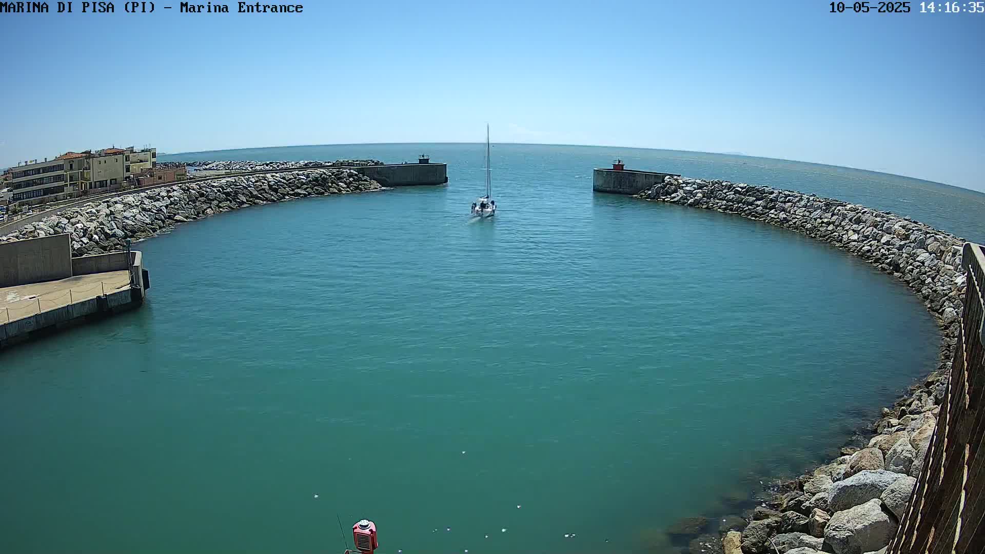 A sailboat navigates a calm, teal harbor enclosed by rocky breakwaters under a clear blue sky.