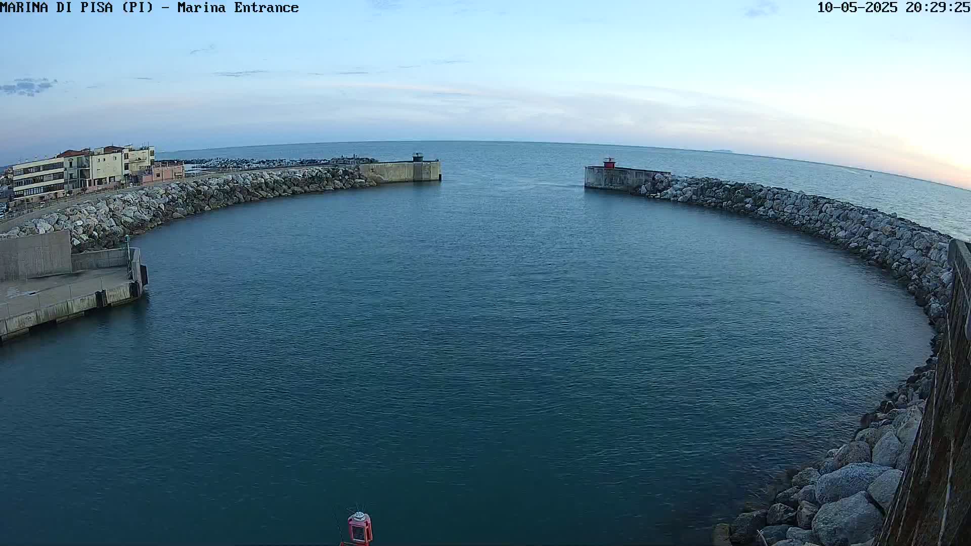 A calm, dark-blue body of water is enclosed by a curved rock wall leading to a harbor entrance with a building visible in the distance under a clear, twilight sky.