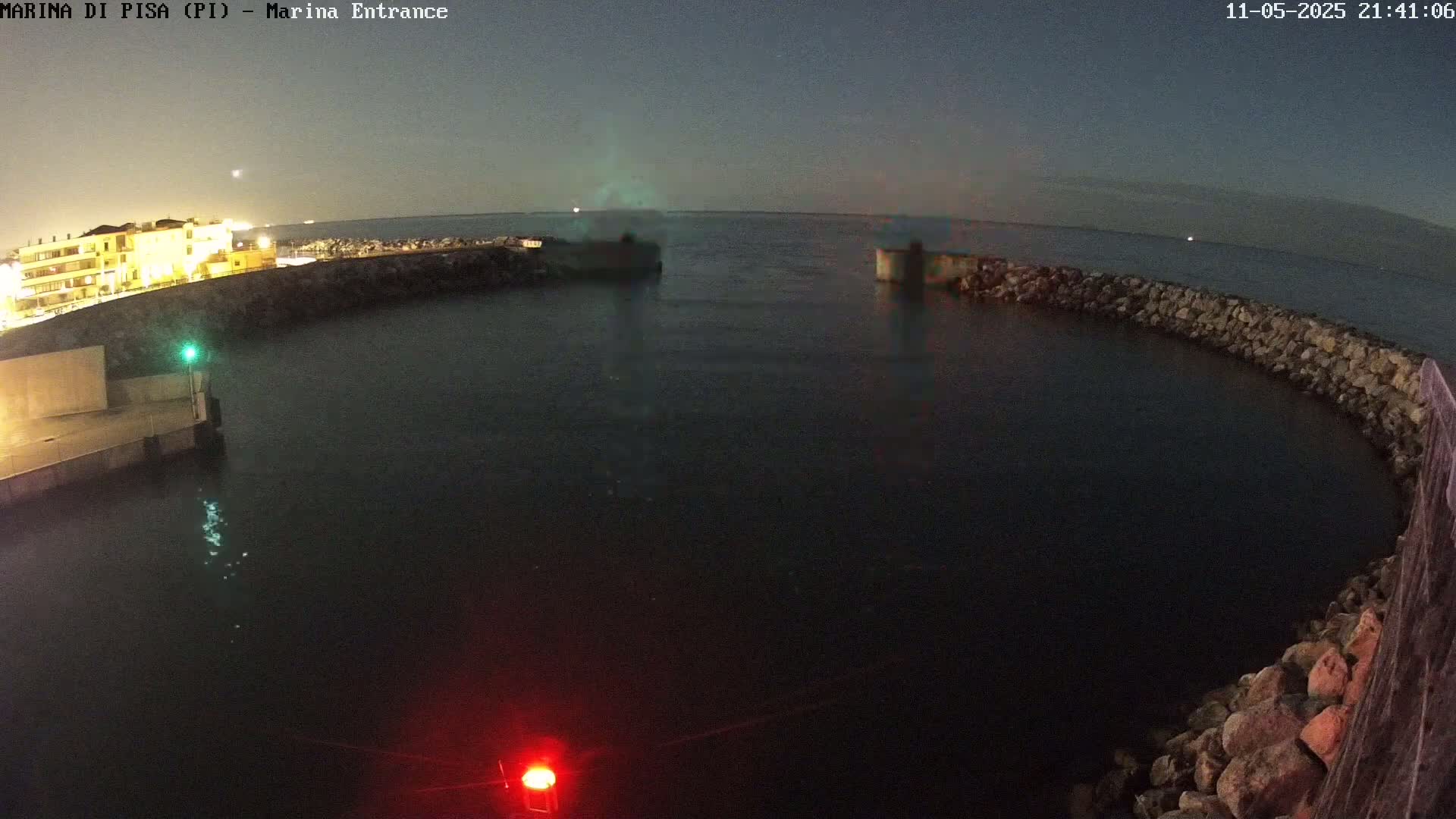A nighttime view of a calm harbor, enclosed by a low stone wall, with a building illuminated on the left and dark water in the center.