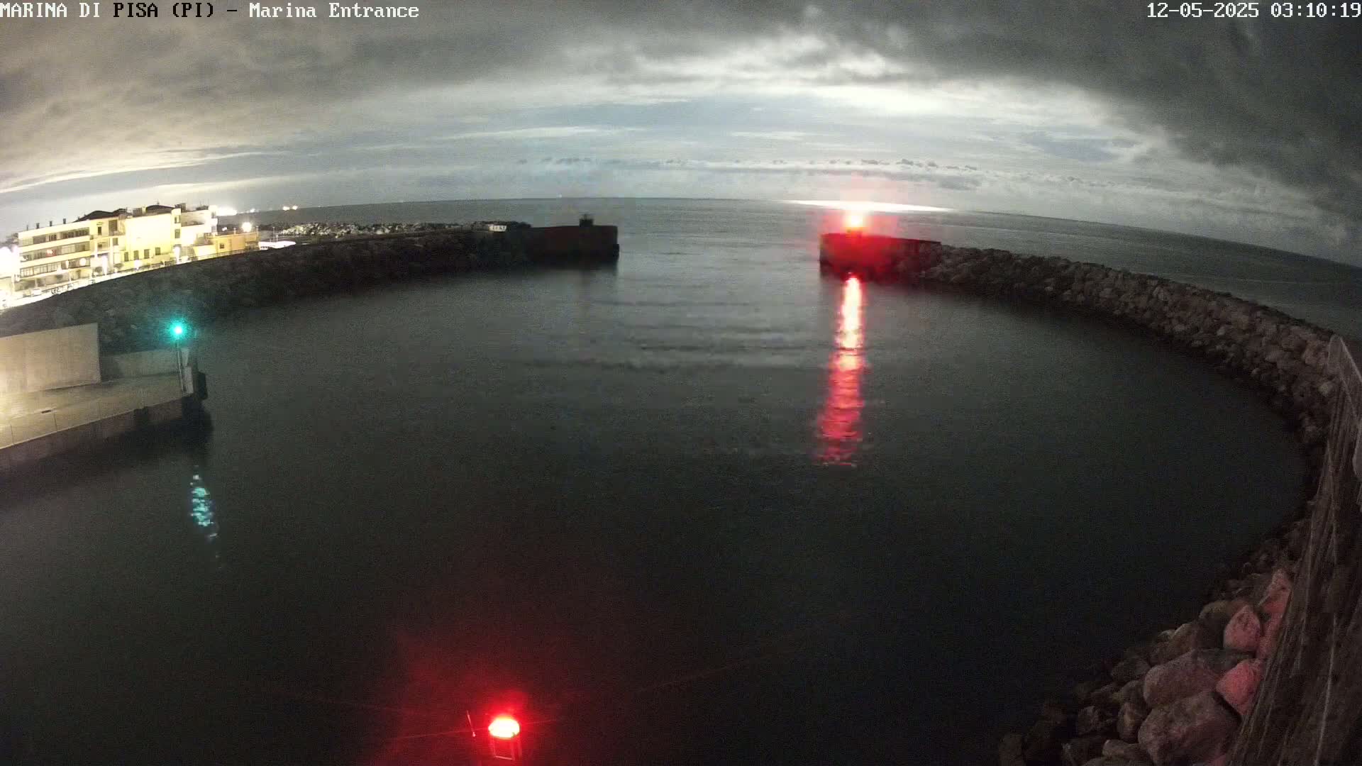 A nighttime view of a harbor entrance with calm water under an overcast sky, featuring red and green navigation lights.