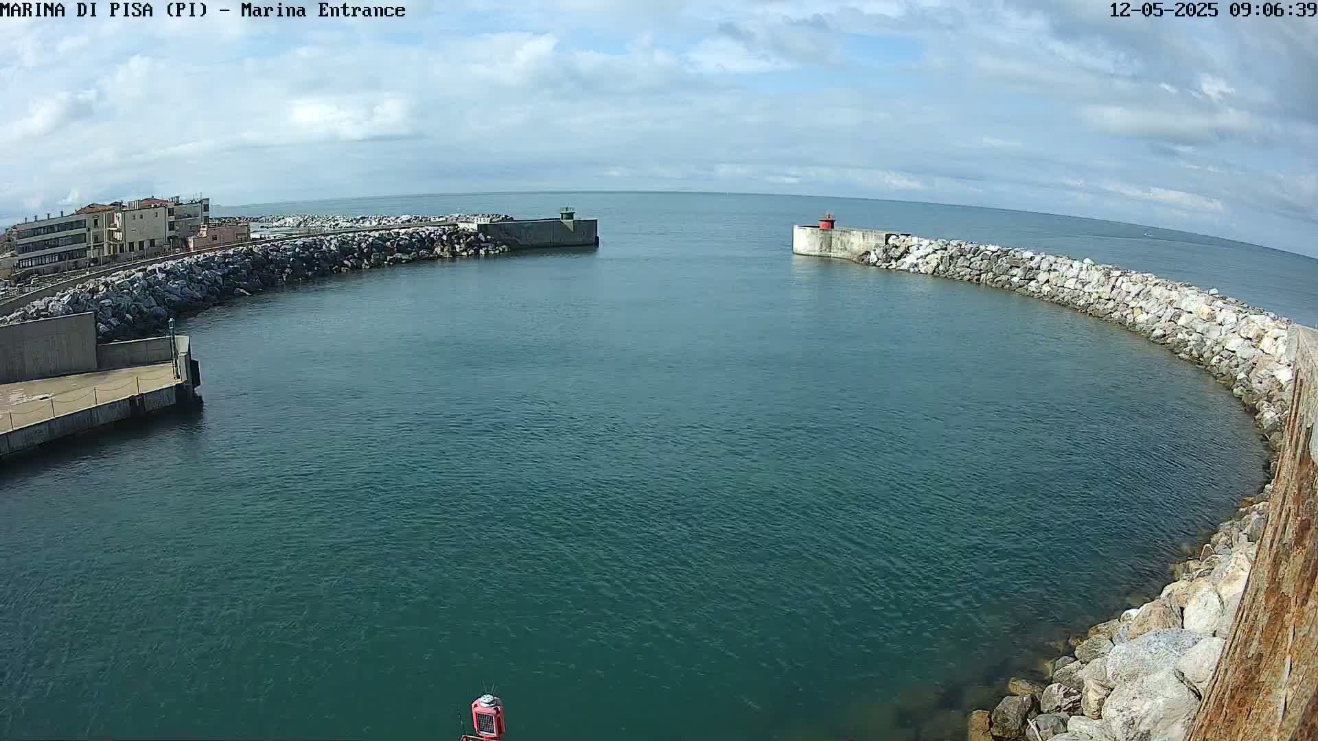 A harbor entrance with calm, teal water is framed by rocky breakwaters under a partly cloudy sky.