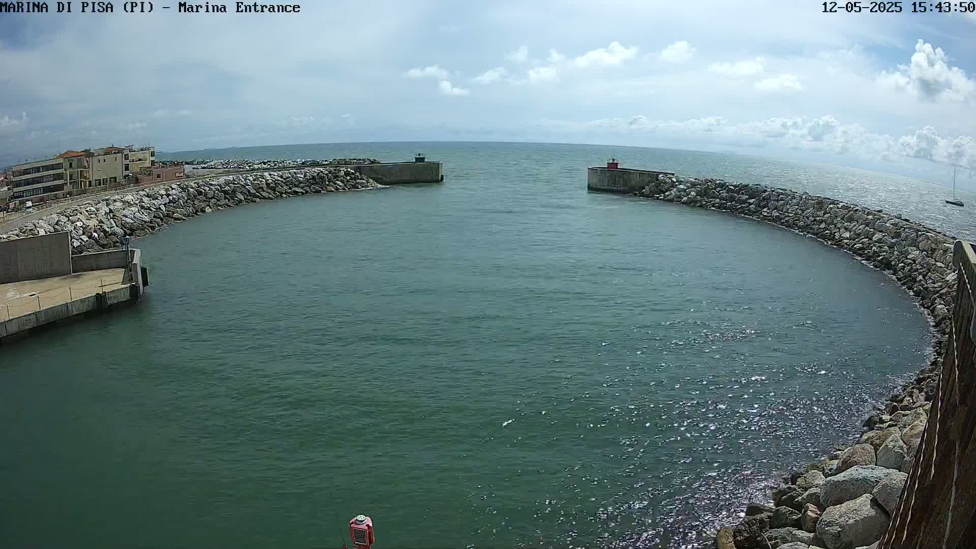 A partly cloudy day shows a harbor entrance with calm, dark-green water enclosed by rock walls and a building visible in the distance.
