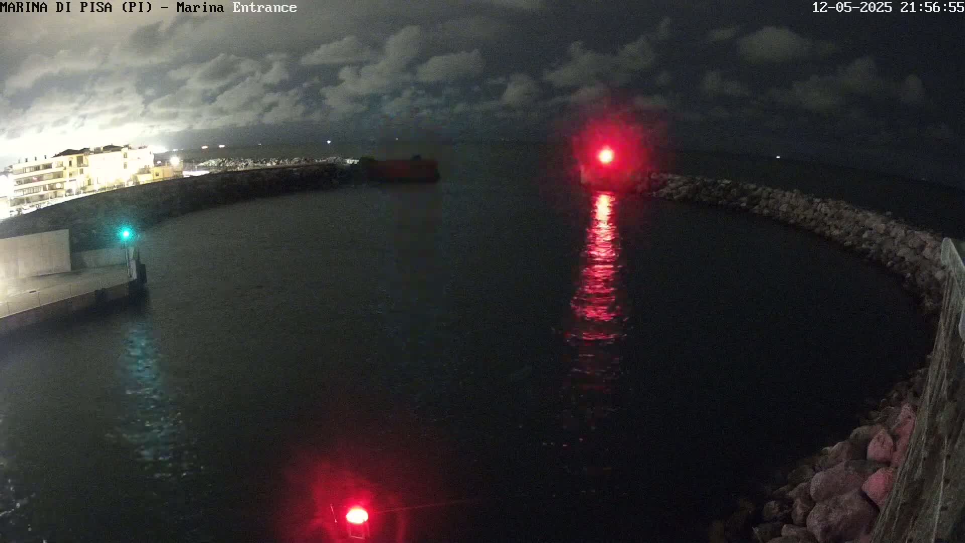 A nighttime view of a harbor entrance with calm water, illuminated by a red light and buildings on the shore under a partly cloudy sky.