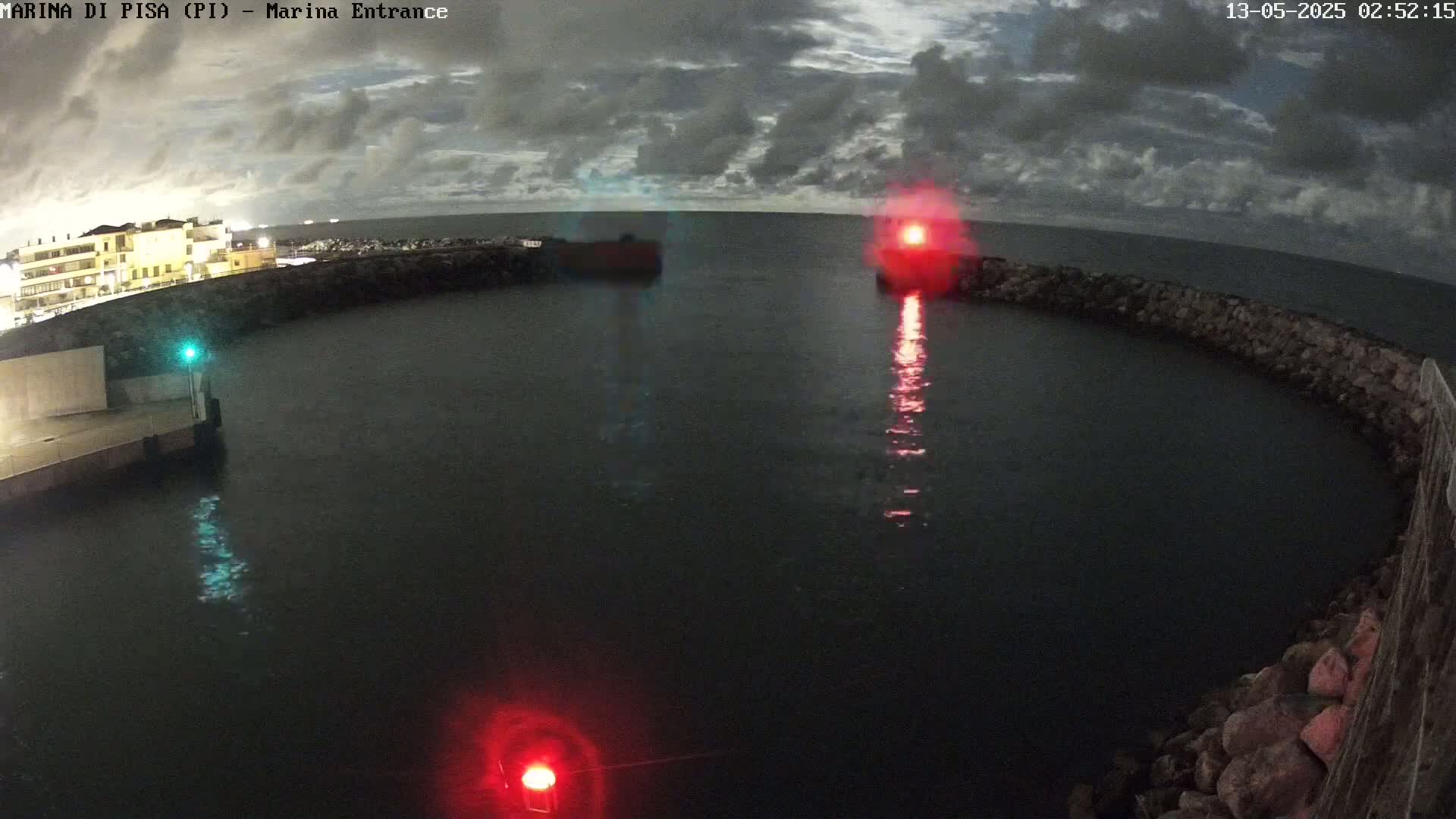 Under a partly cloudy night sky, a dark body of water is enclosed by a low stone wall, with red and green lights visible at the entrance.