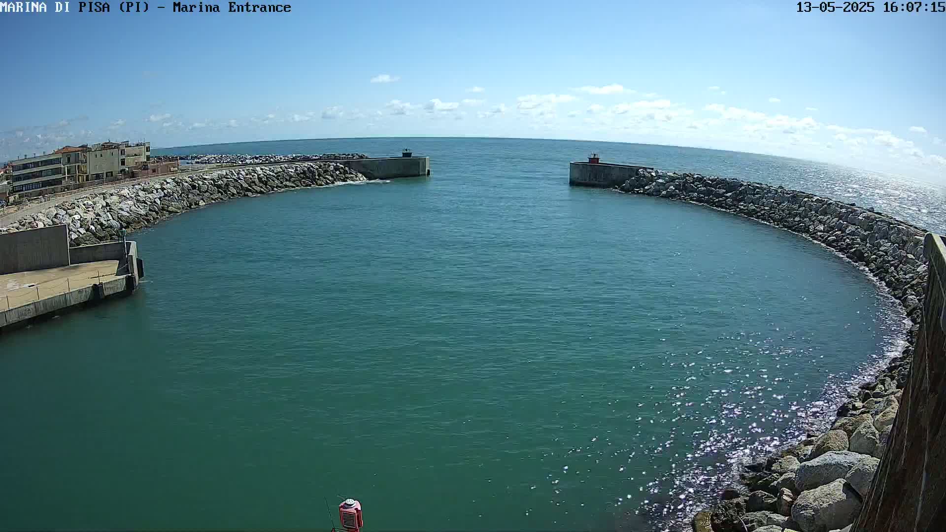 A marina entrance with calm, teal water is bordered by rocky breakwaters under a mostly sunny sky.