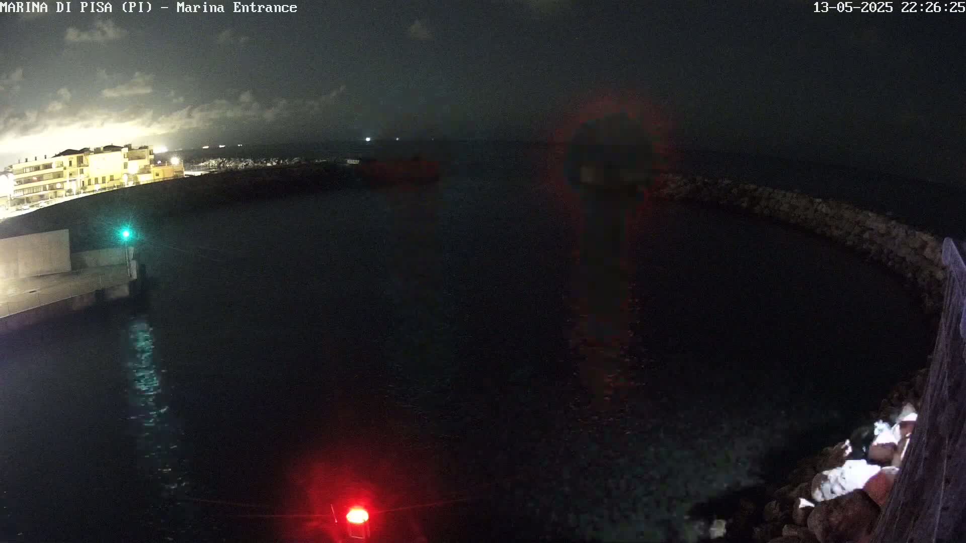 A nighttime view of a dark harbor with a stone wall, illuminated buildings in the distance, and a few lights visible on the water.