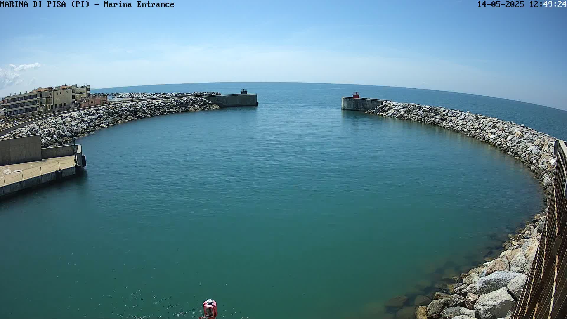 A harbor entrance with calm, teal water is bordered by rock and concrete walls under a clear blue sky.