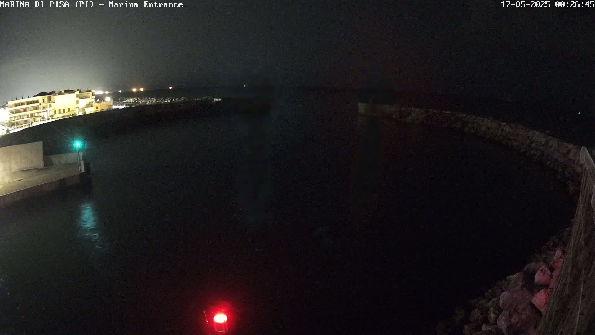 A nighttime, dark view of a marina entrance with calm water, a building faintly visible in the distance, and a stone wall curving into the water.
