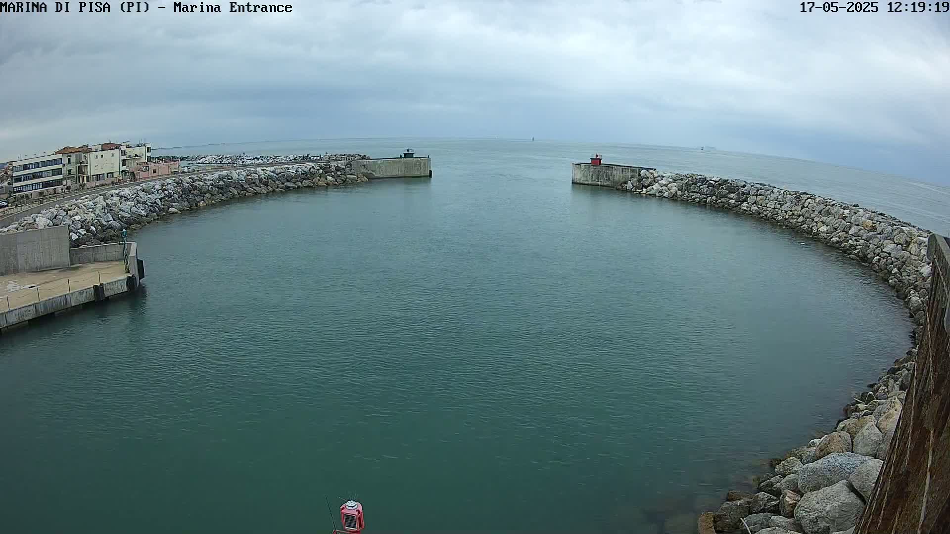 A harbor entrance with calm, dark-green water under an overcast sky is framed by stone walls and a building in the background.