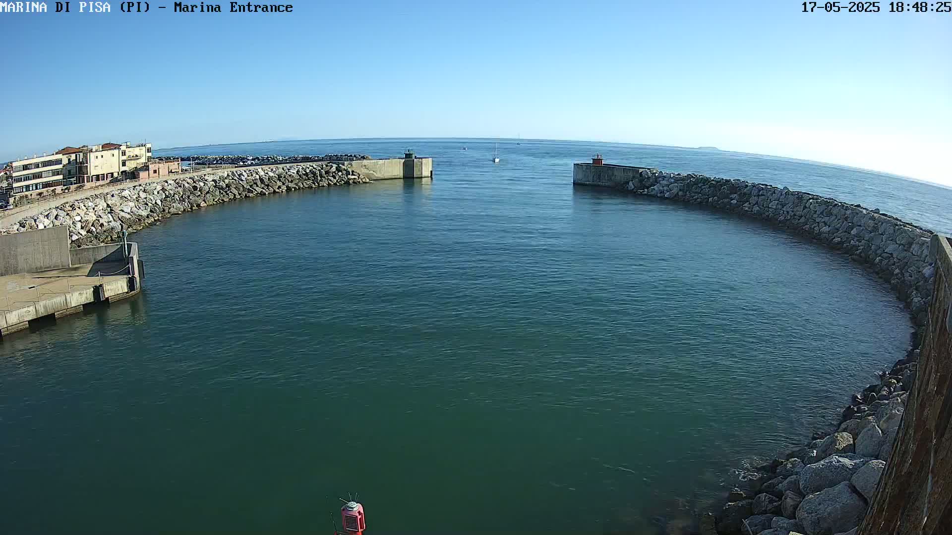 A calm, clear day shows a harbor entrance with stone walls and a small sailboat in the distance.