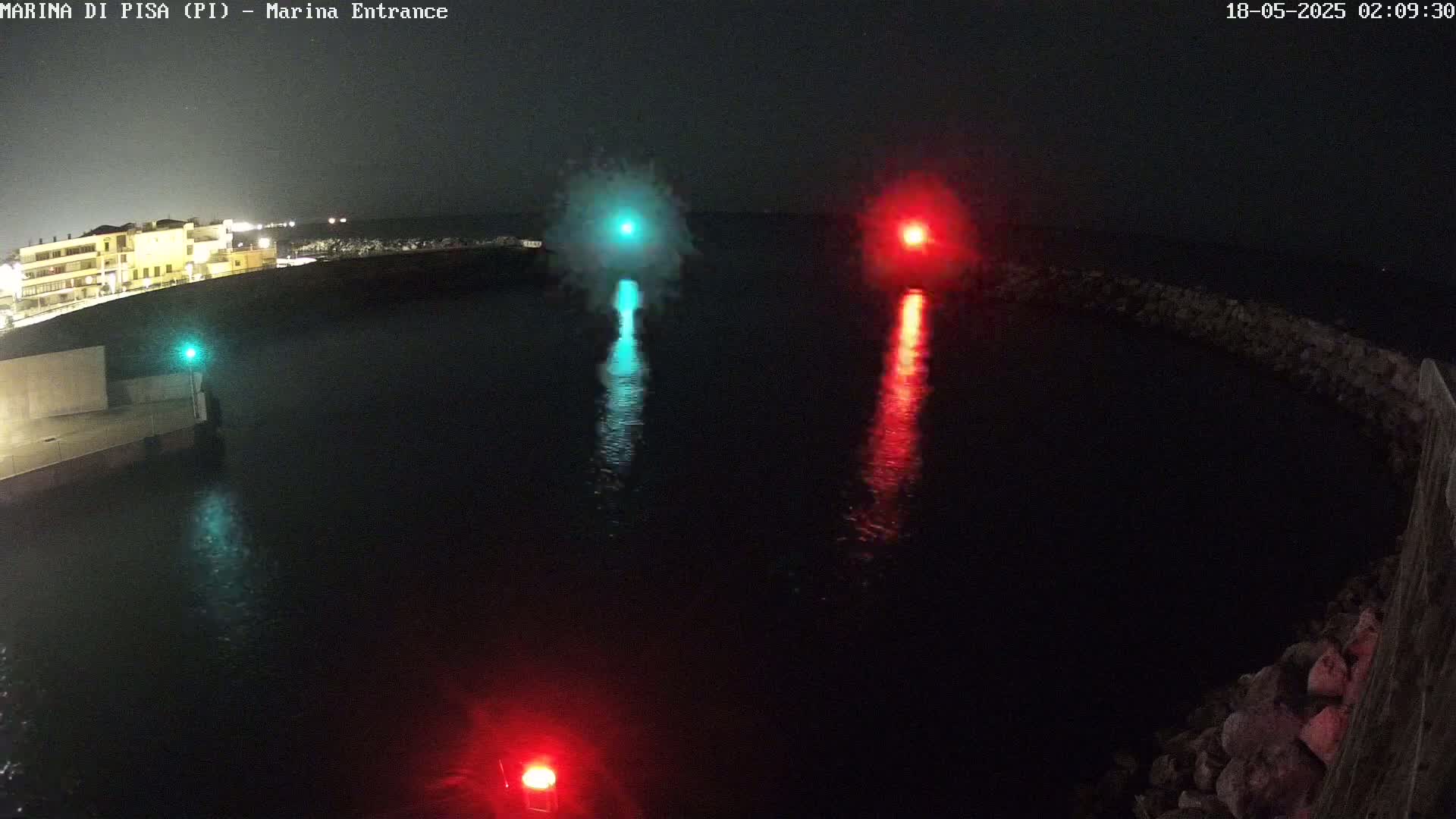 A nighttime harbor scene shows a teal and red light reflecting on dark water, with a building faintly visible in the background.