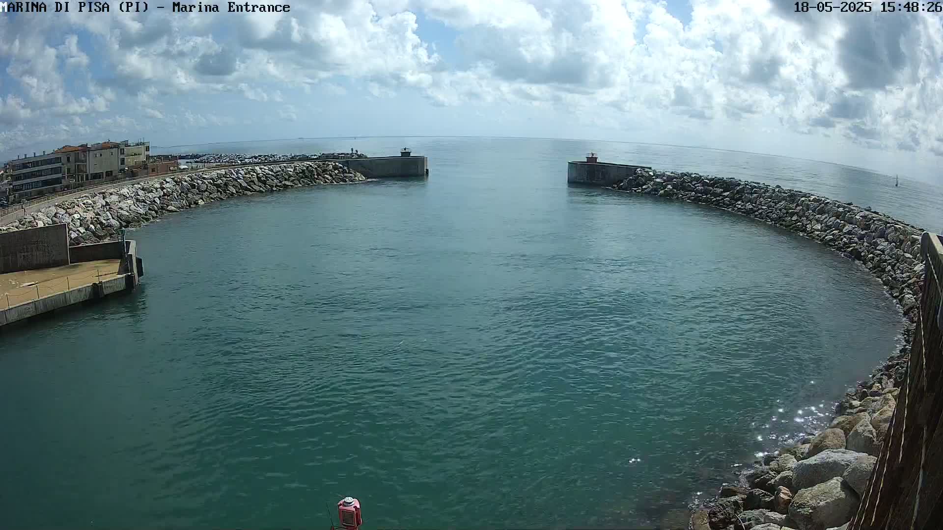 A harbor entrance with calm, teal water is partially enclosed by a curved rock wall under a partly cloudy sky.