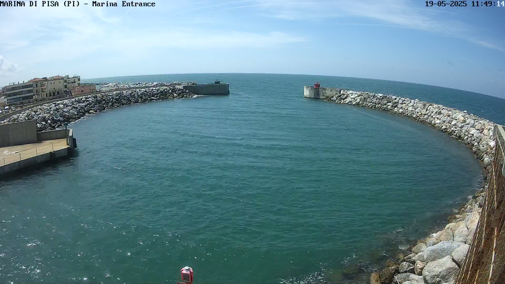 A harbor entrance with calm, teal water is surrounded by stone walls, and buildings are visible in the background under a mostly sunny sky.