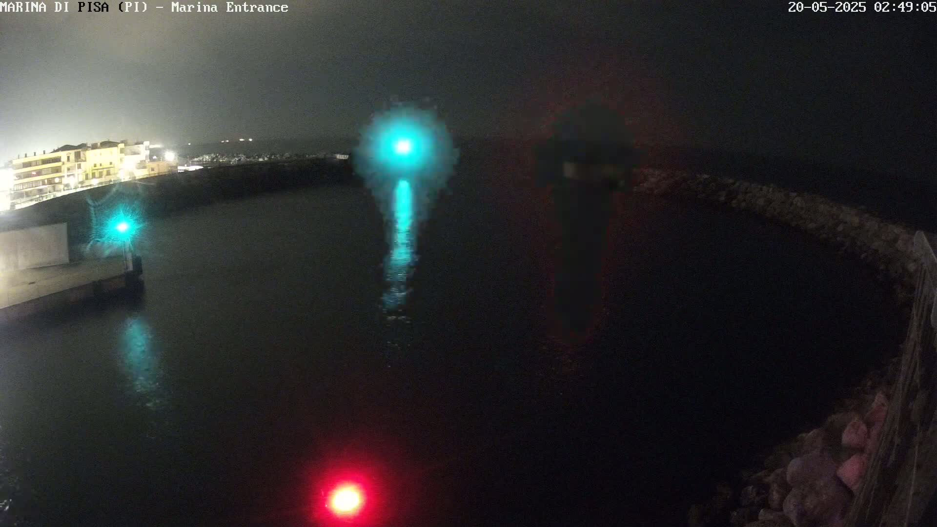 A nighttime view of a dark harbor with a teal and red light visible on the water, and a building illuminated in the background under a dark, cloudy sky.