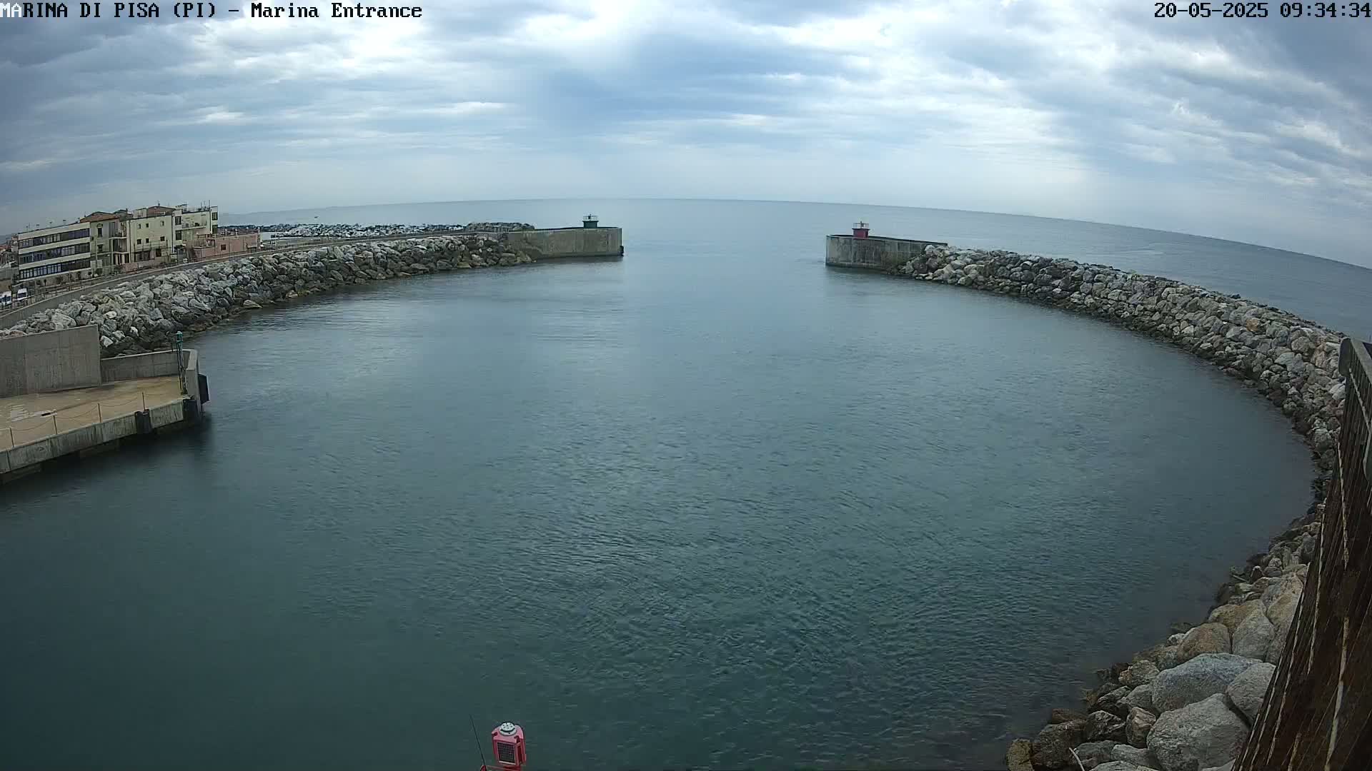 A harbor entrance with calm, dark-blue water is partially enclosed by stone walls under a cloudy sky.