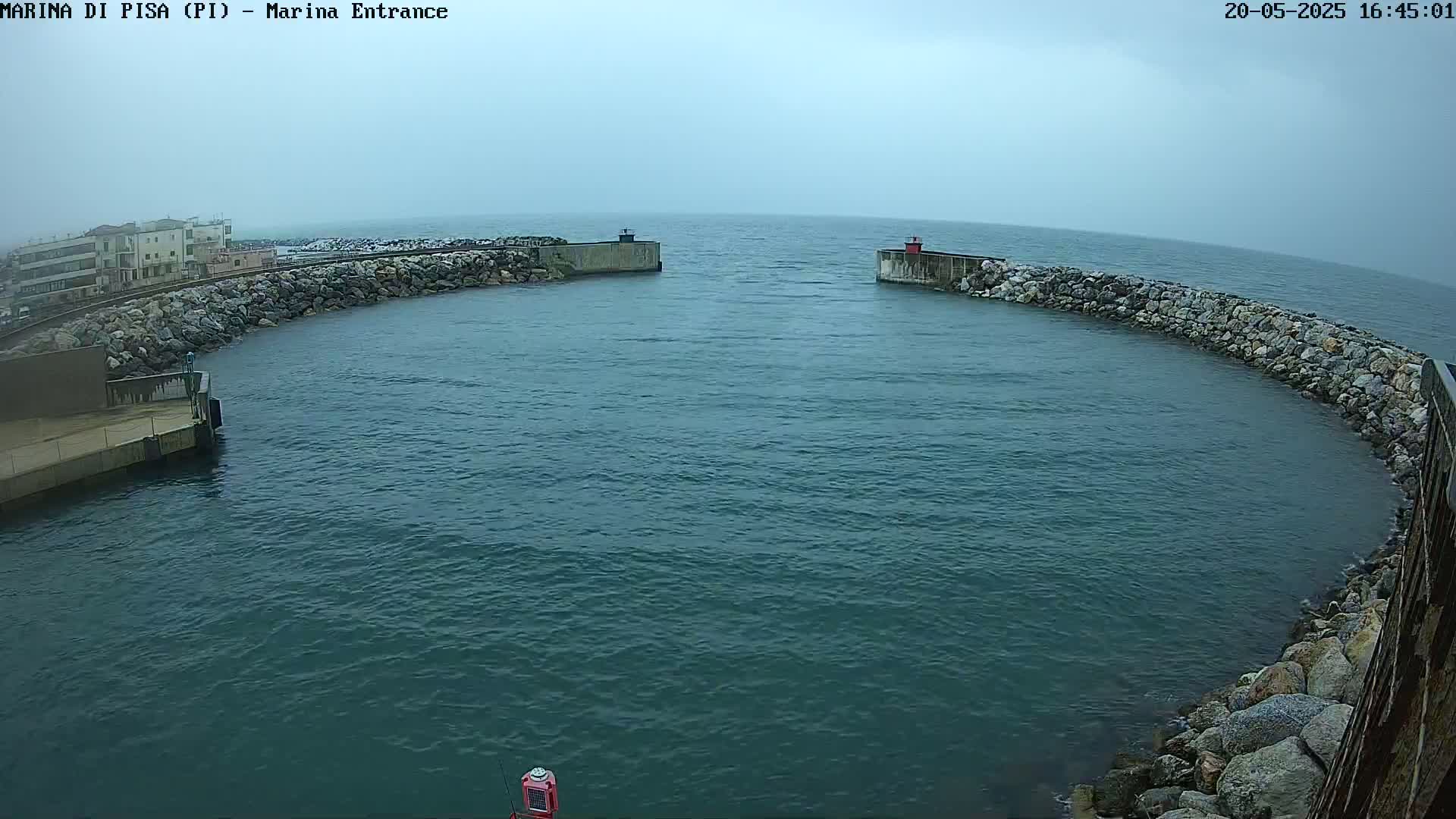 A harbor entrance with calm, dark-blue water under an overcast sky is framed by rock walls and a building in the distance.