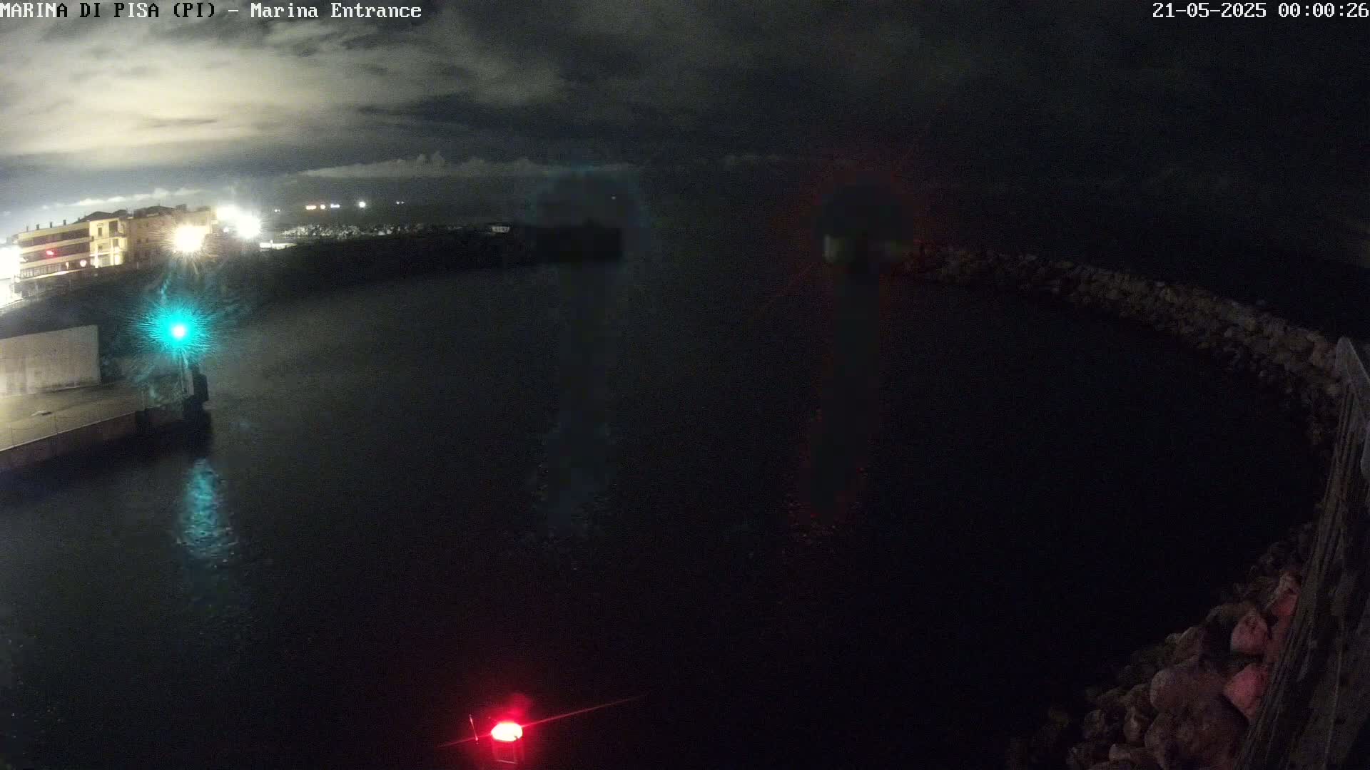 A nighttime view of a dark harbor entrance with a few lights visible on the buildings across the water under a cloudy sky.