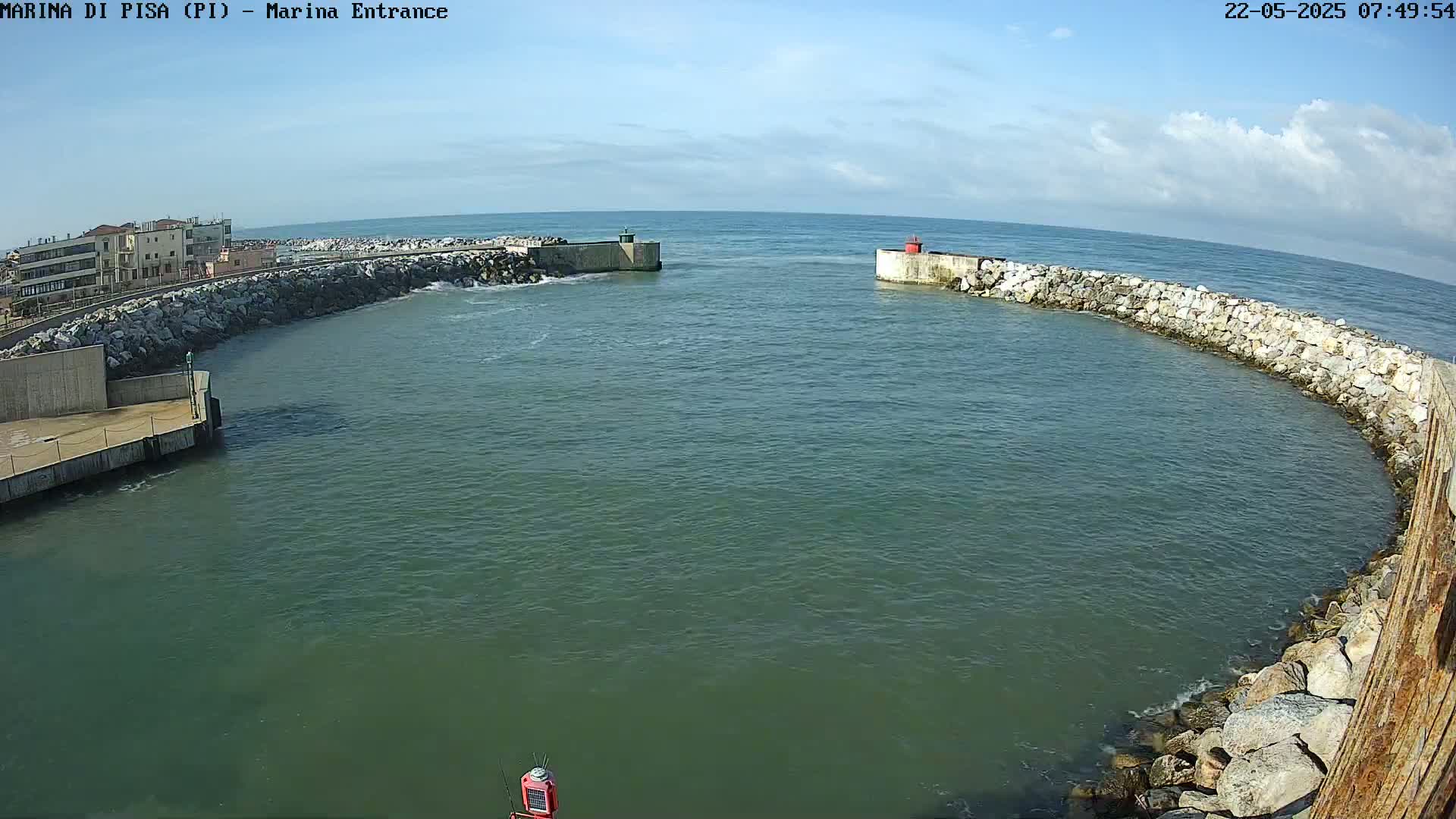 A partly cloudy day shows a harbor entrance with calm water, stone walls, and buildings in the background.