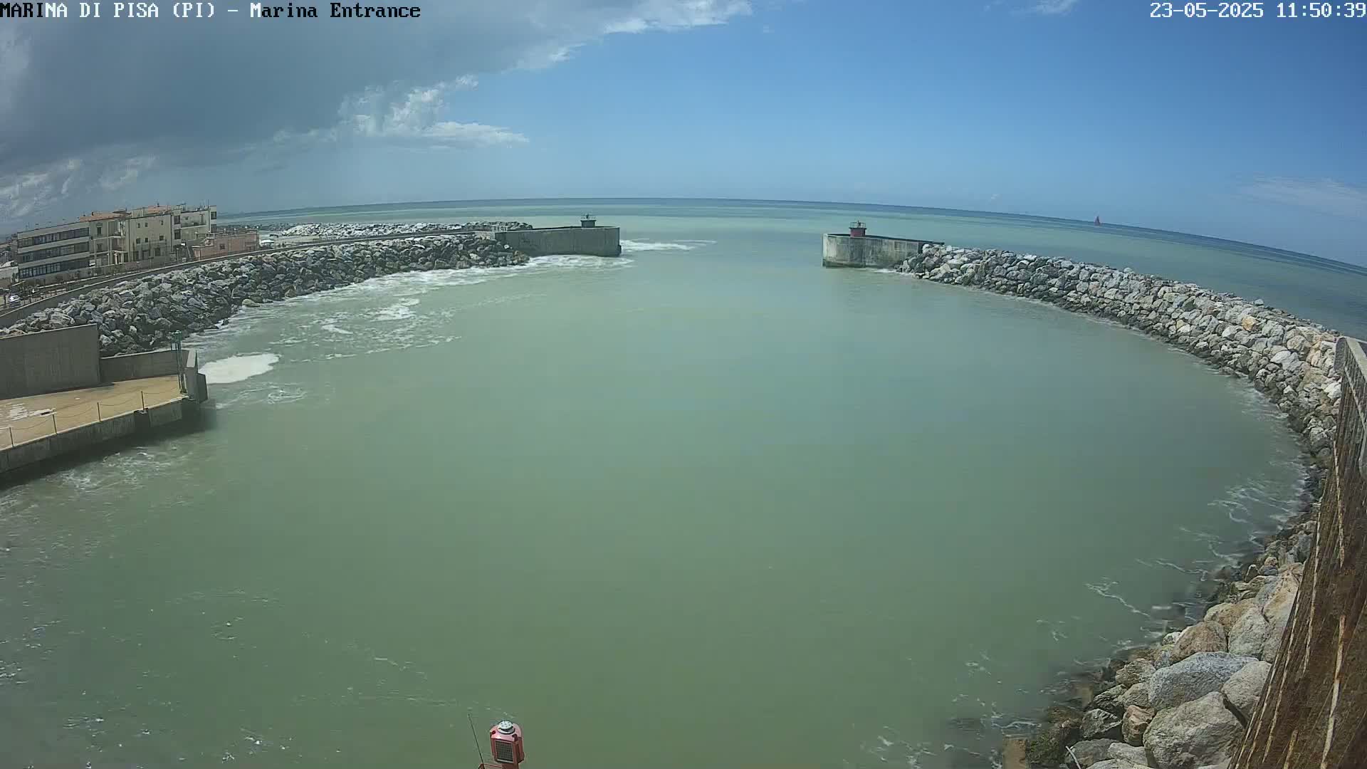 A harbor entrance with calm, greenish water under a mostly sunny sky, partially enclosed by rock walls and bordered by buildings.