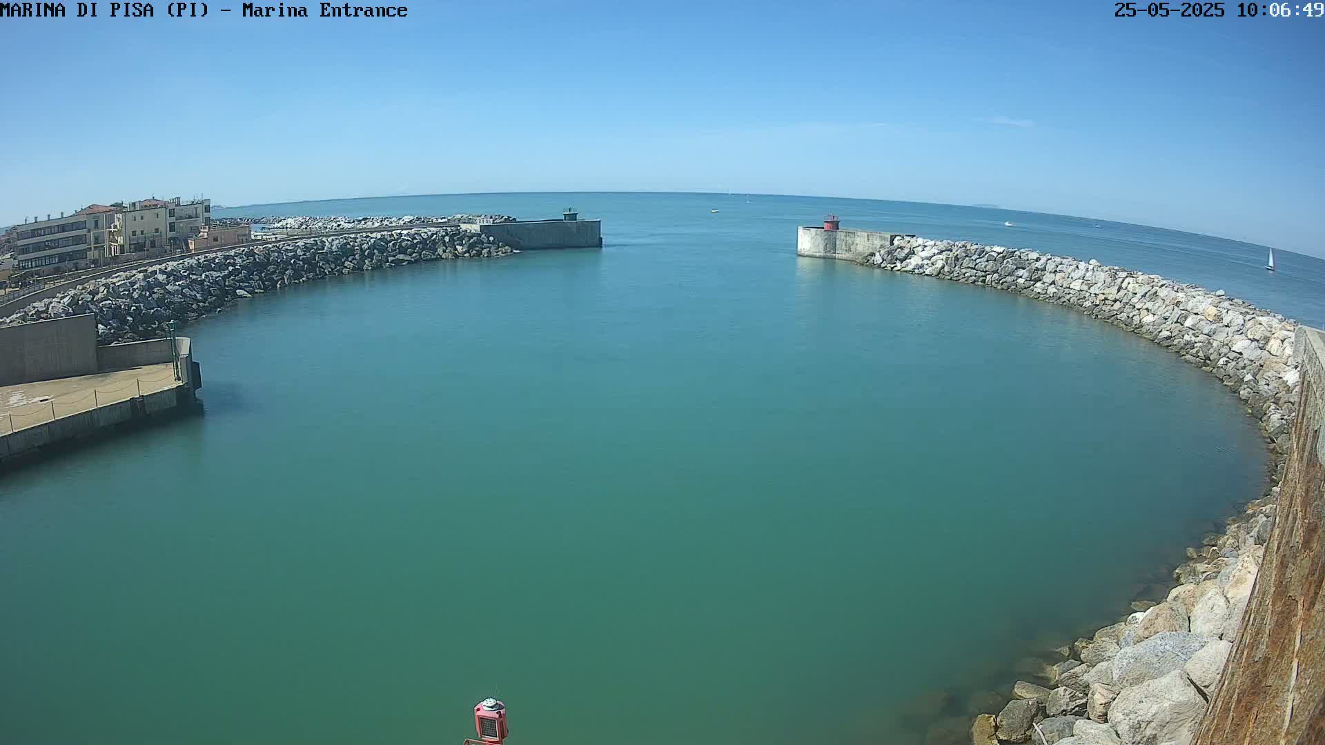 A harbor entrance with calm, teal water is bordered by rock walls and a building on the left, under a clear blue sky.