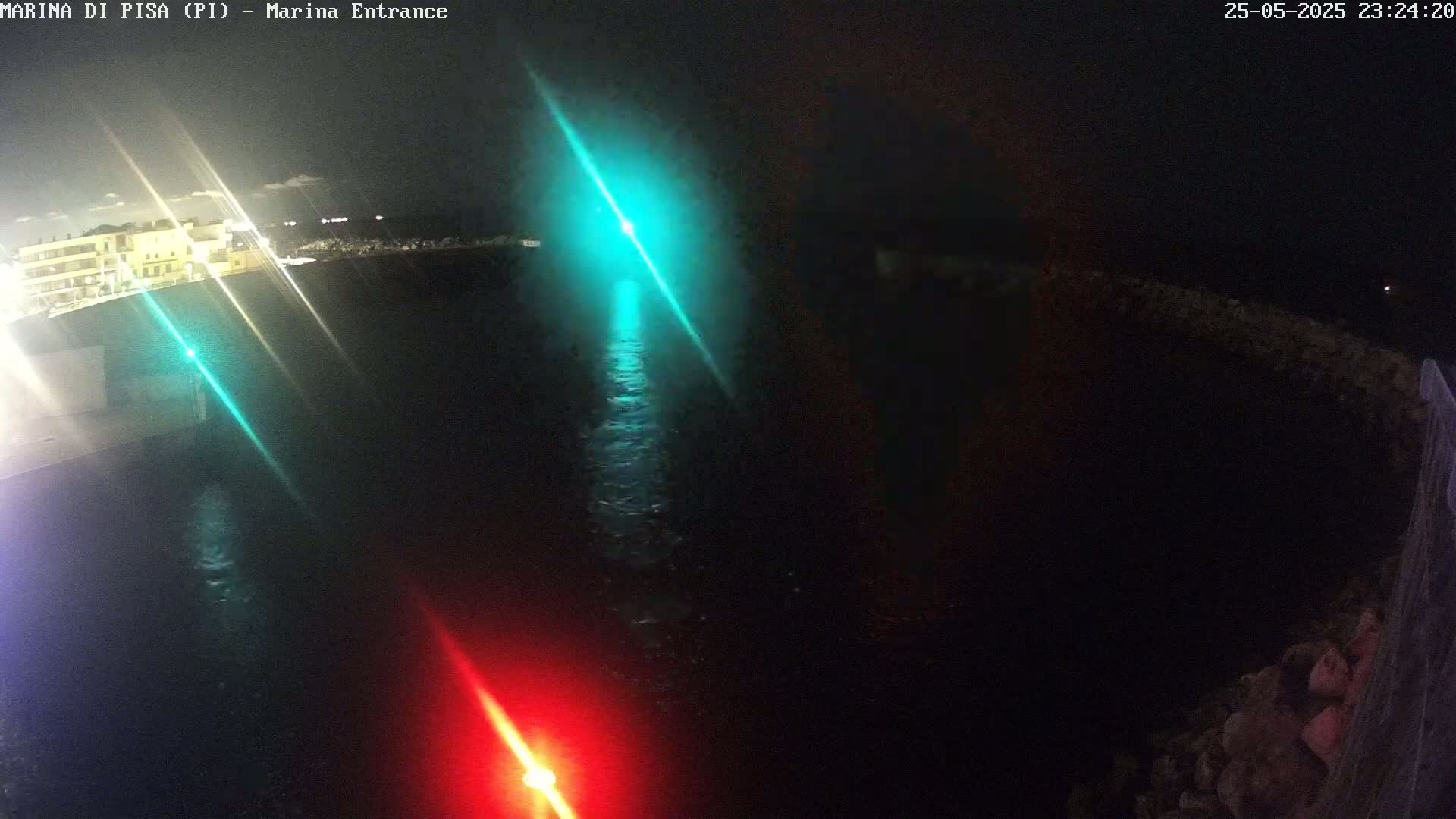 A nighttime aerial view shows a dark body of water illuminated by green and red lights emanating from either side of a channel, with a building visible in the background.