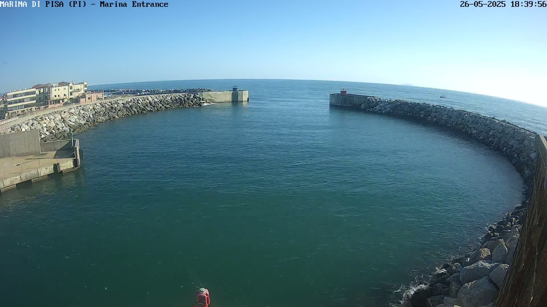 A harbor entrance with calm, clear water is nestled between two rock-lined breakwaters on a sunny day.