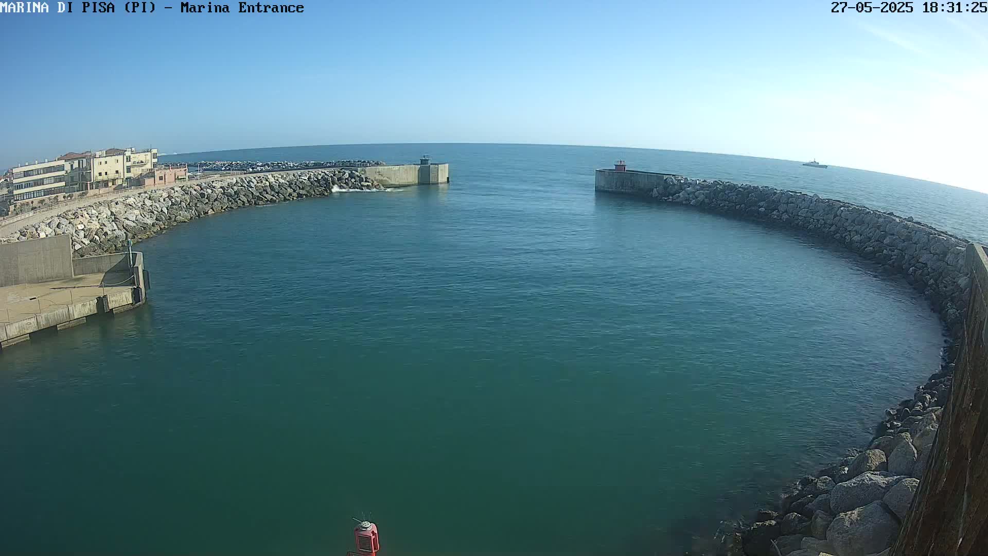 A marina entrance with calm, teal water is seen under a clear blue sky, enclosed by rock walls and a building in the background.