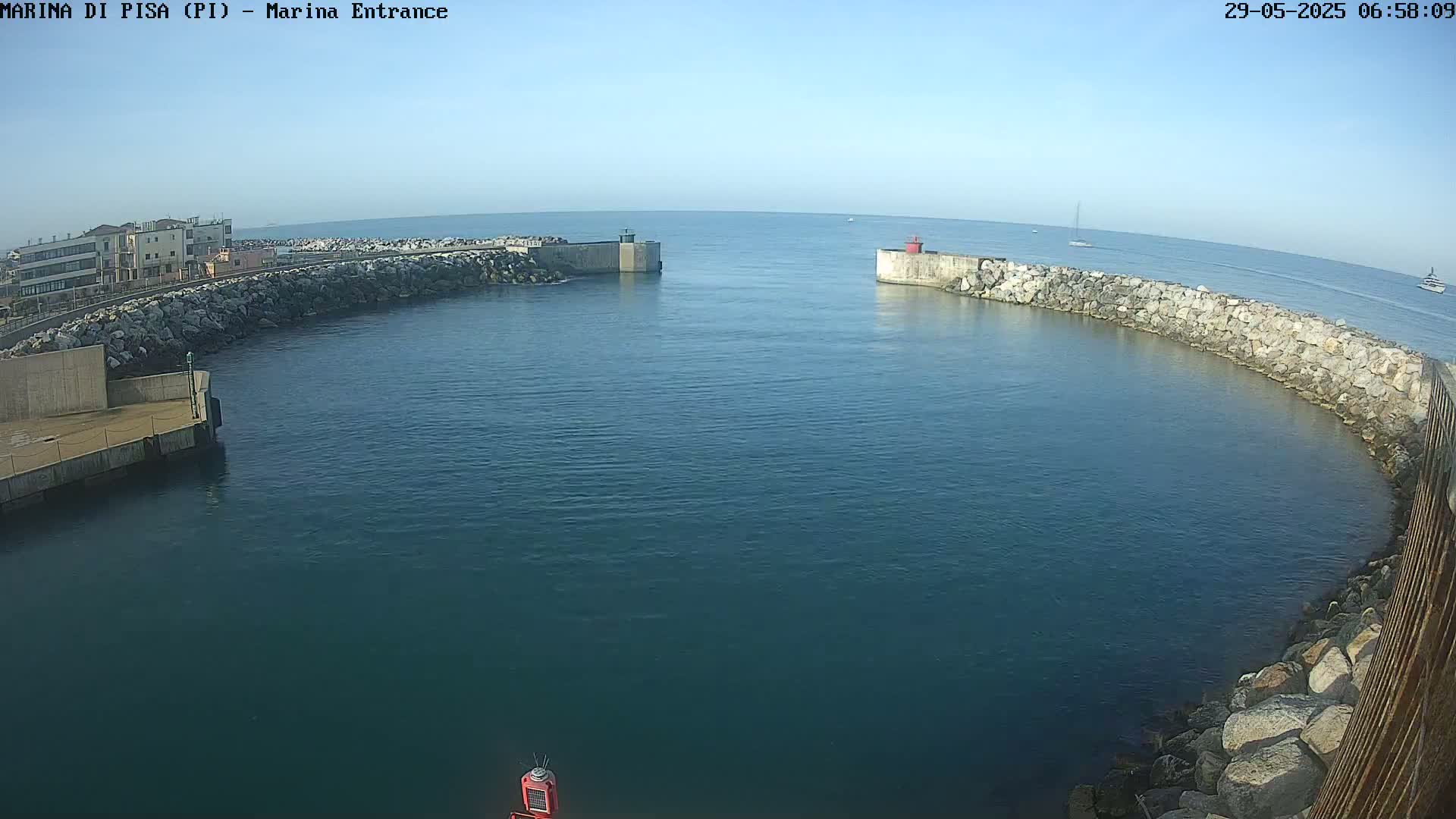 A calm, clear day reveals a harbor entrance, defined by stone walls enclosing a tranquil body of dark blue water, with a few distant boats visible on the horizon.